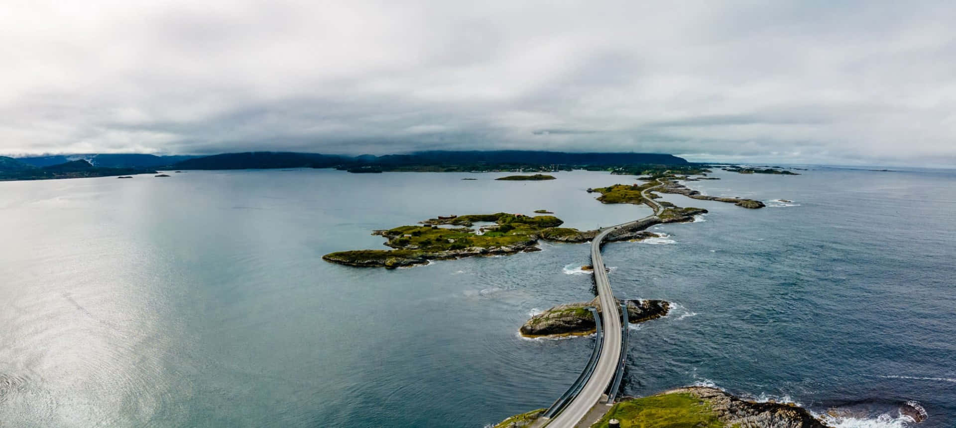 Road 桌布 壁紙 — 一條蜿蜒的海岸公路在陰天之下，蜿蜒穿過岩石島嶼和碧綠的海水。