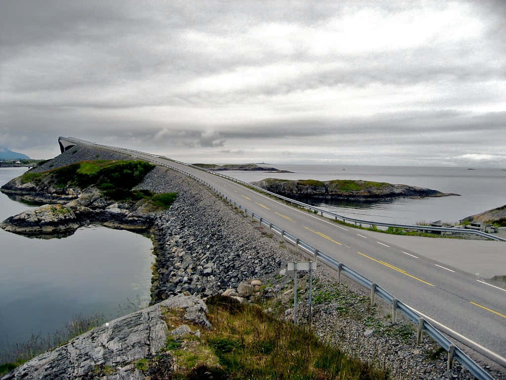 Road 桌布 壁紙 — 一條蜿蜒的海岸公路在陰天之下蜿蜒穿過岩石島嶼，下方是平靜的水面。