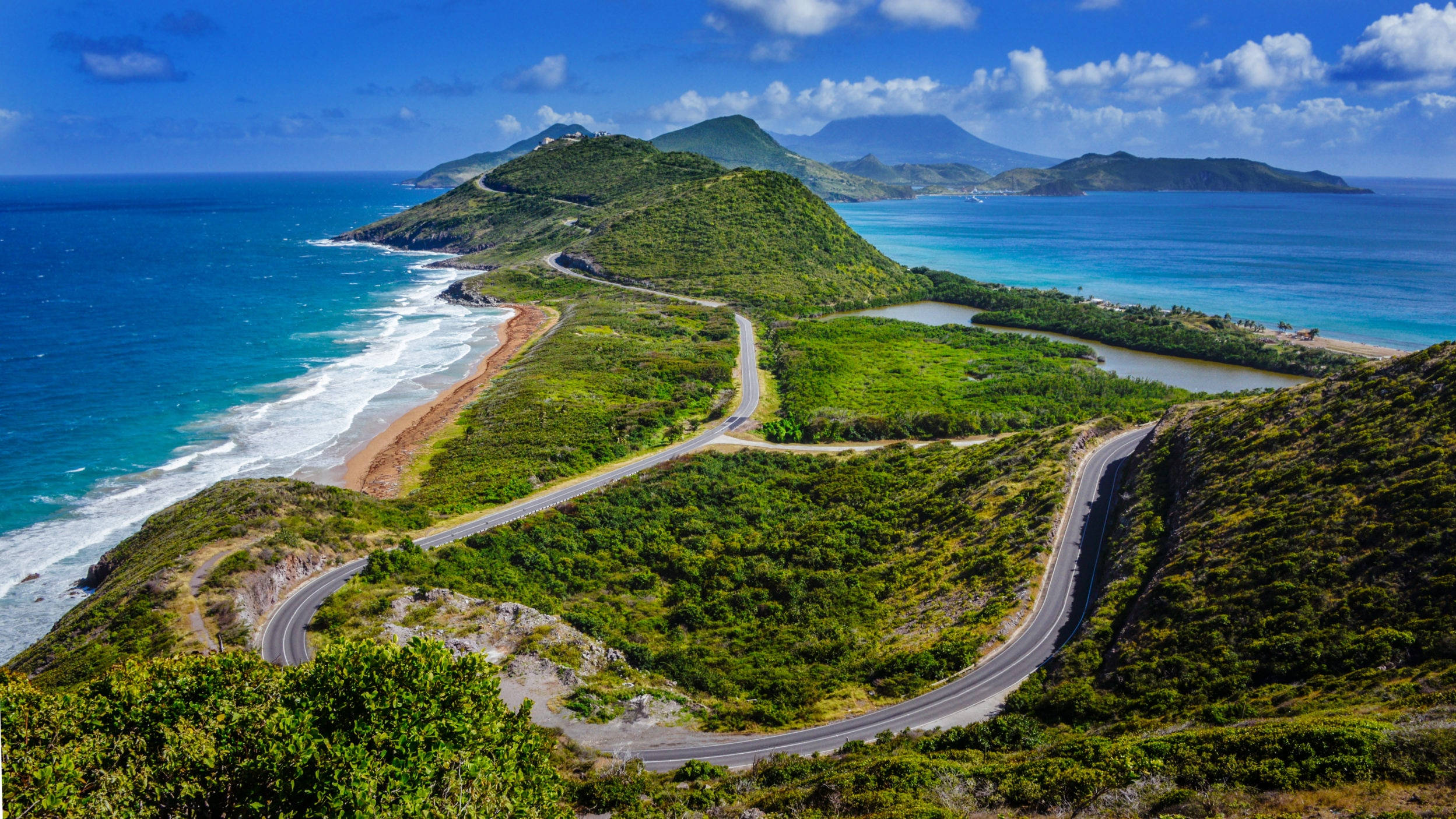 島嶼 桌布 壁紙 — 一條蜿蜒的海岸公路穿過鬱鬱蔥蔥的綠色山丘，旁邊是碧綠的海水，上方是明亮的藍天和蓬鬆的白雲。