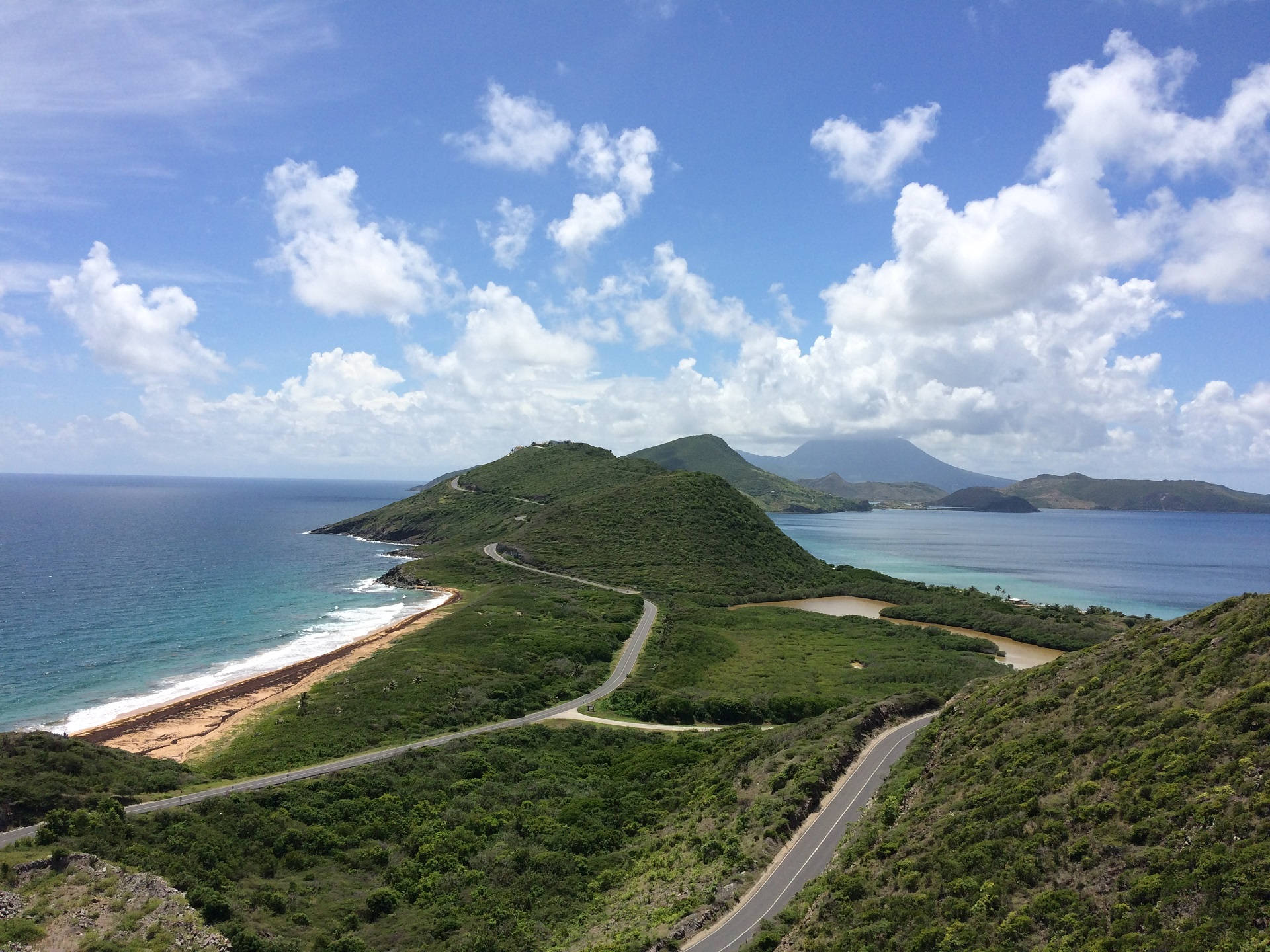 島嶼 桌布 壁紙 — 一條蜿蜒的海岸公路穿過鬱鬱蔥蔥的綠色山丘，上方是明亮的藍天和蓬鬆的白雲。