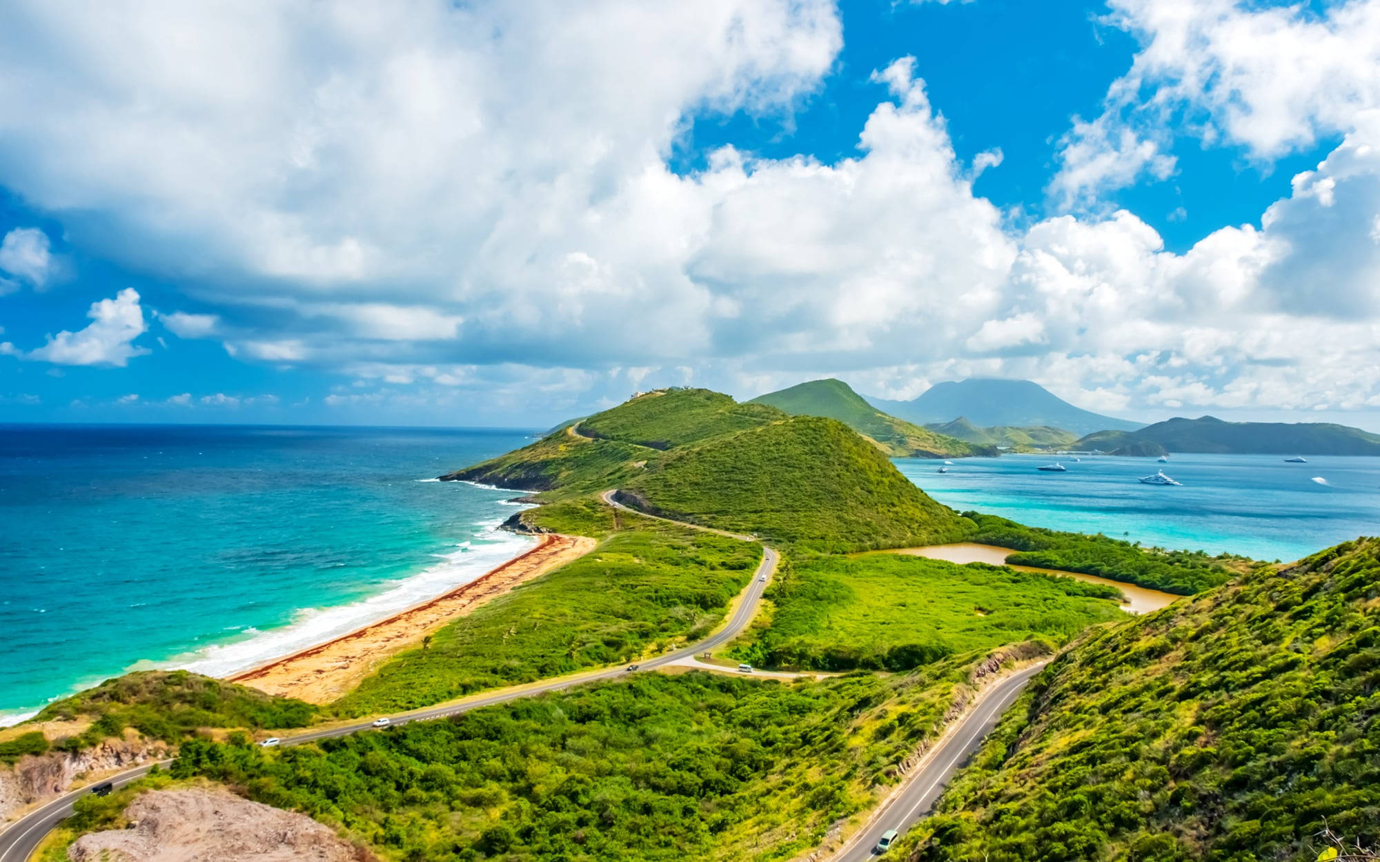 Caribbean 桌布 壁紙 — 一條蜿蜒的海岸公路緊貼著翠綠的山丘，旁邊是碧綠的海水，上方是明亮的藍天和蓬鬆的白雲。