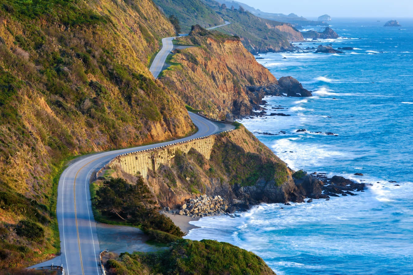 highway 桌布 壁紙 — 一條蜿蜒的海岸公路緊貼陡峭的懸崖，俯瞰著碧綠的海浪拍打岩石海岸。