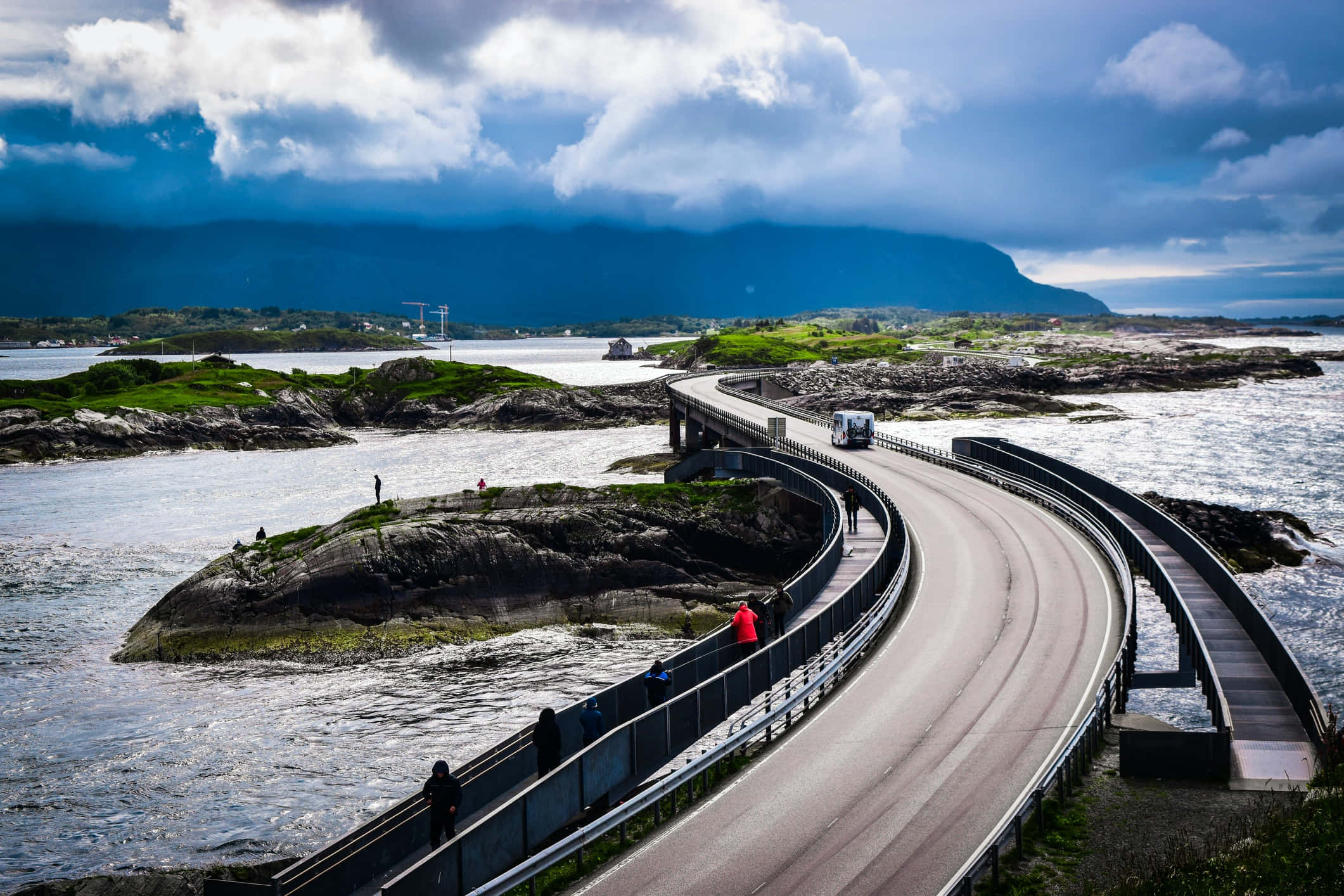 Road 桌布 壁紙 — 一條蜿蜒的海岸道路優雅地彎曲在岩石島嶼上，人們在行走，一輛貨車在戲劇性的多雲天空下行駛。