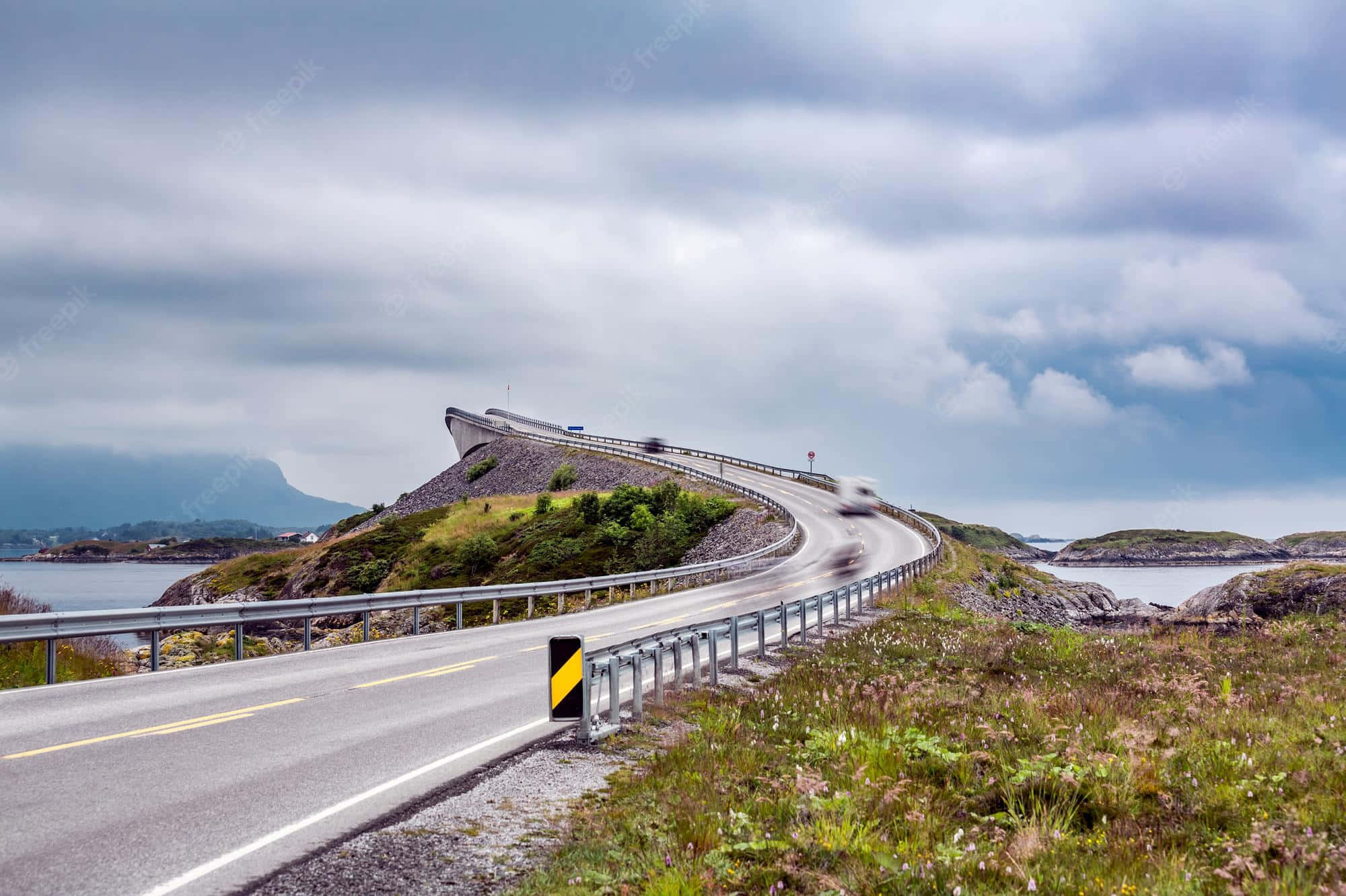 Road 桌布 壁紙 — 一條蜿蜒的海岸道路在岩石地形上戲劇性地彎曲，背景是霧氣籠罩的山脈和水域，天空多雲。