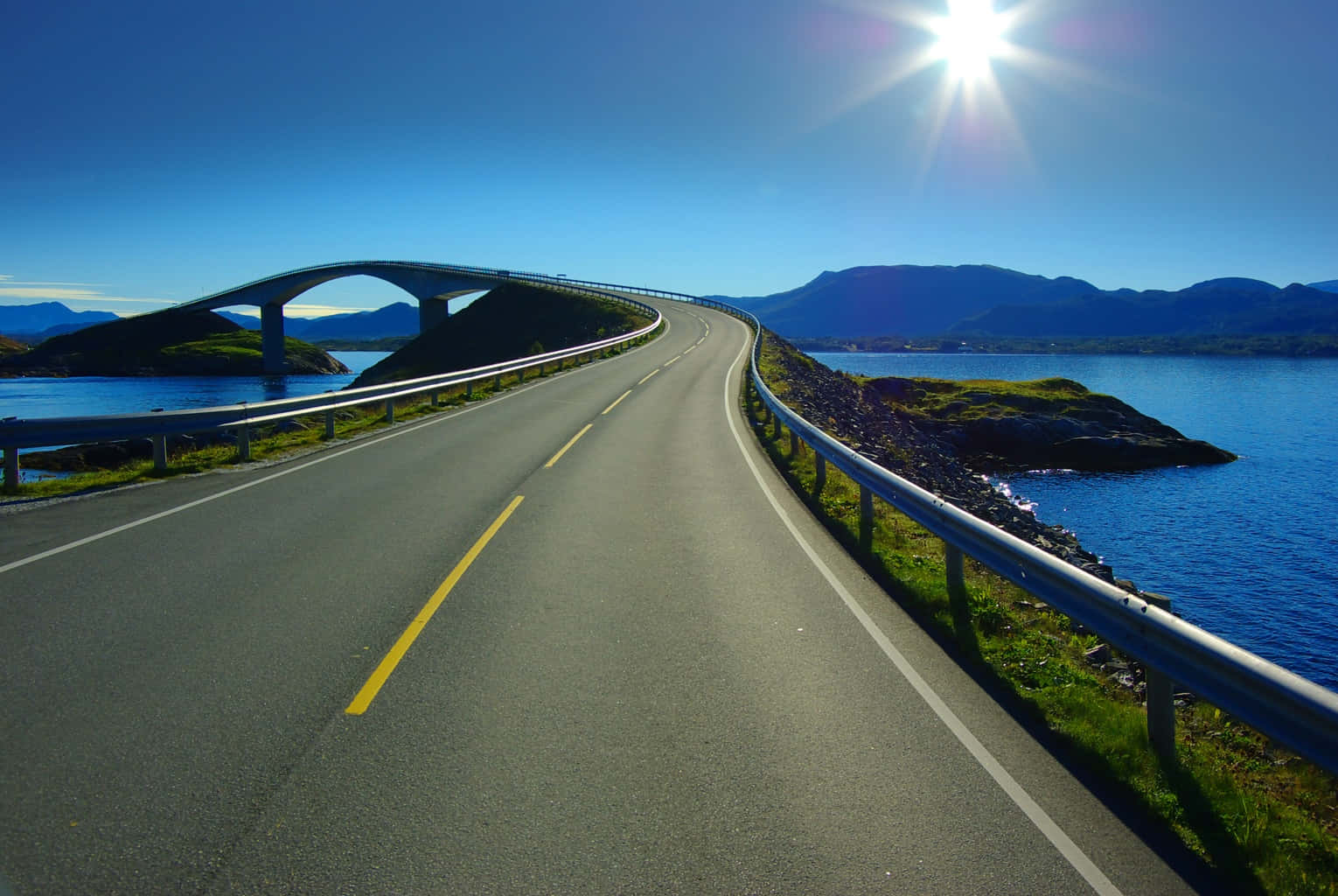 Road 桌布 壁紙 — 一條蜿蜒的海岸道路在明亮的陽光下，越過一座現代橋樑，下方是深藍色的海水和遠處的山脈。