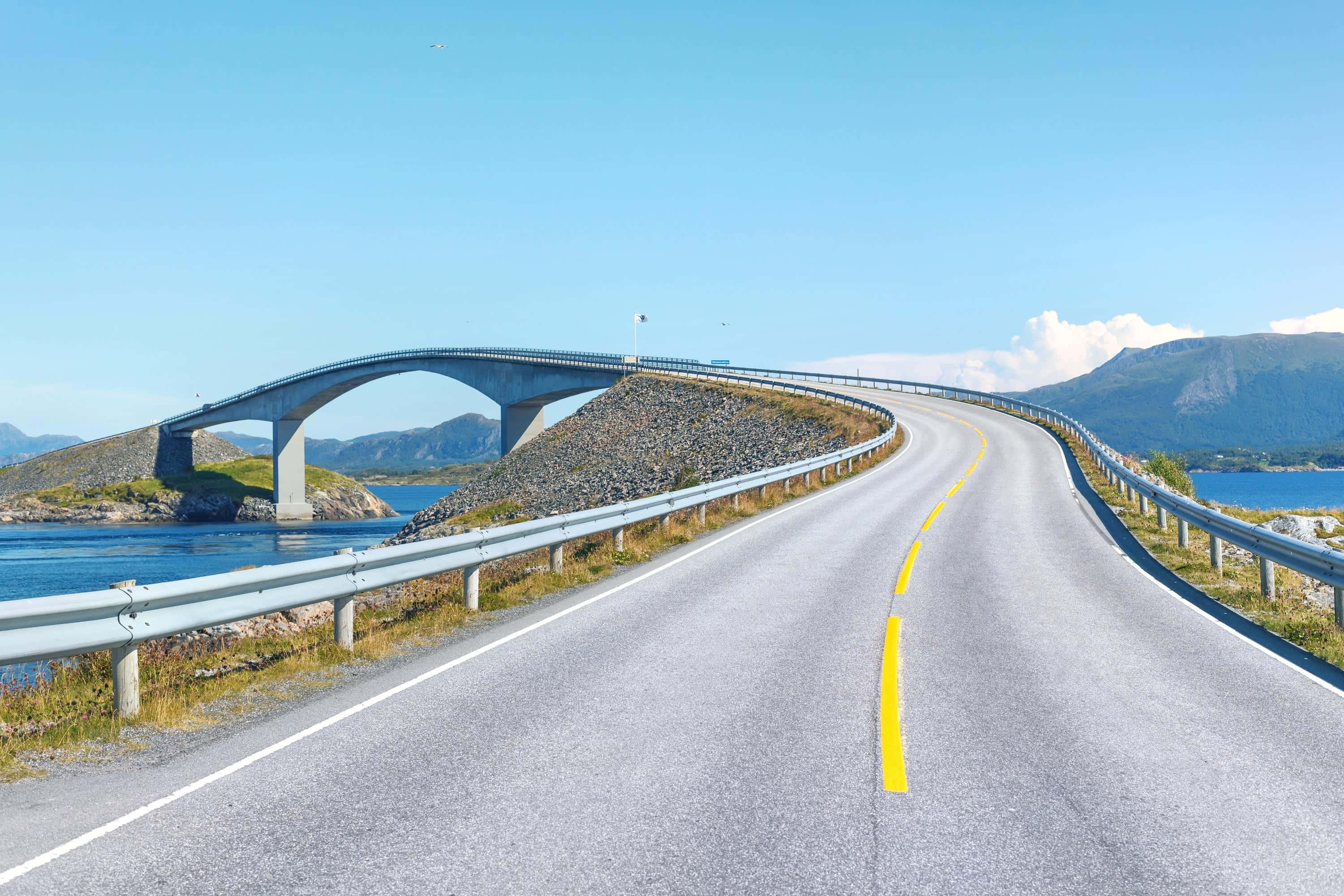 Road 桌布 壁紙 — 一條蜿蜒的海岸道路在現代拱橋下優美地彎曲，背景是山脈和藍色海水。