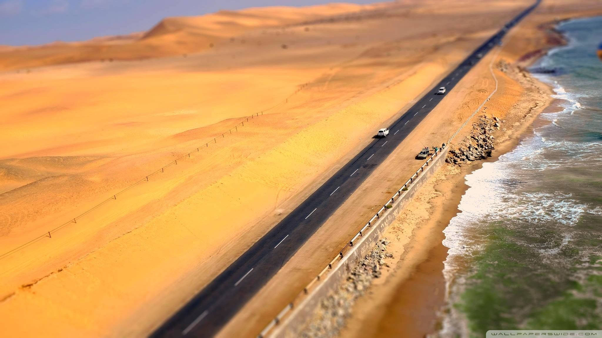 Namibia 桌布 壁紙 — 一條蜿蜒的海岸高速公路穿過金色沙漠沙丘，旁邊是碧綠的海浪，在晴朗的藍天下。