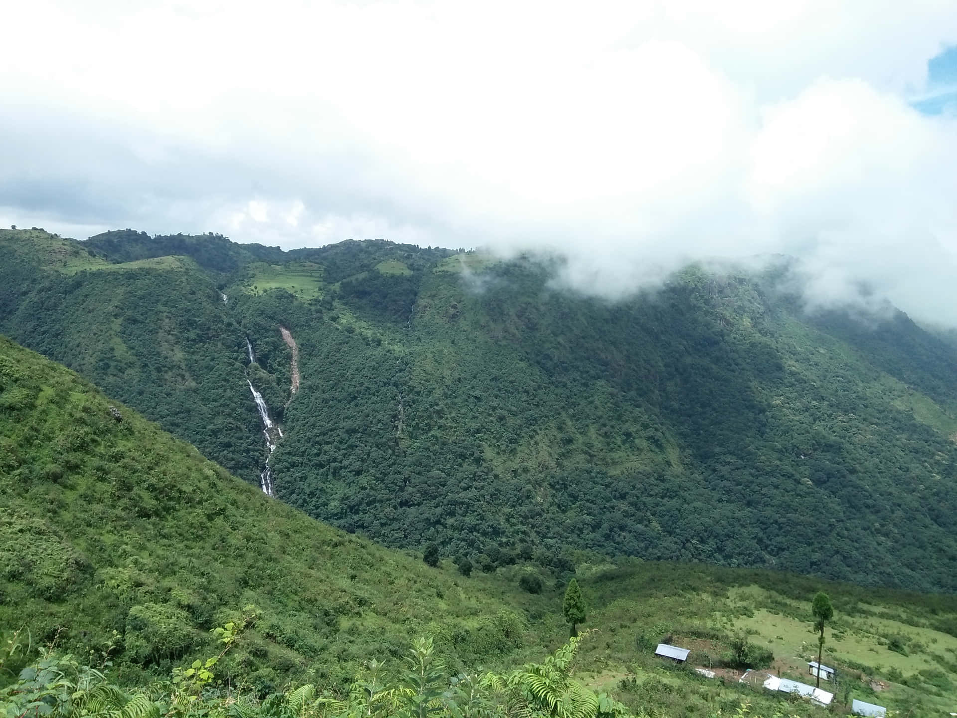 Mountains covered with clouds 壁紙 — 一條蜿蜒的瀑布從鬱鬱蔥蔥的綠色山谷中傾瀉而下，部分被飄動的雲霧遮蓋，下方有一個小村莊。