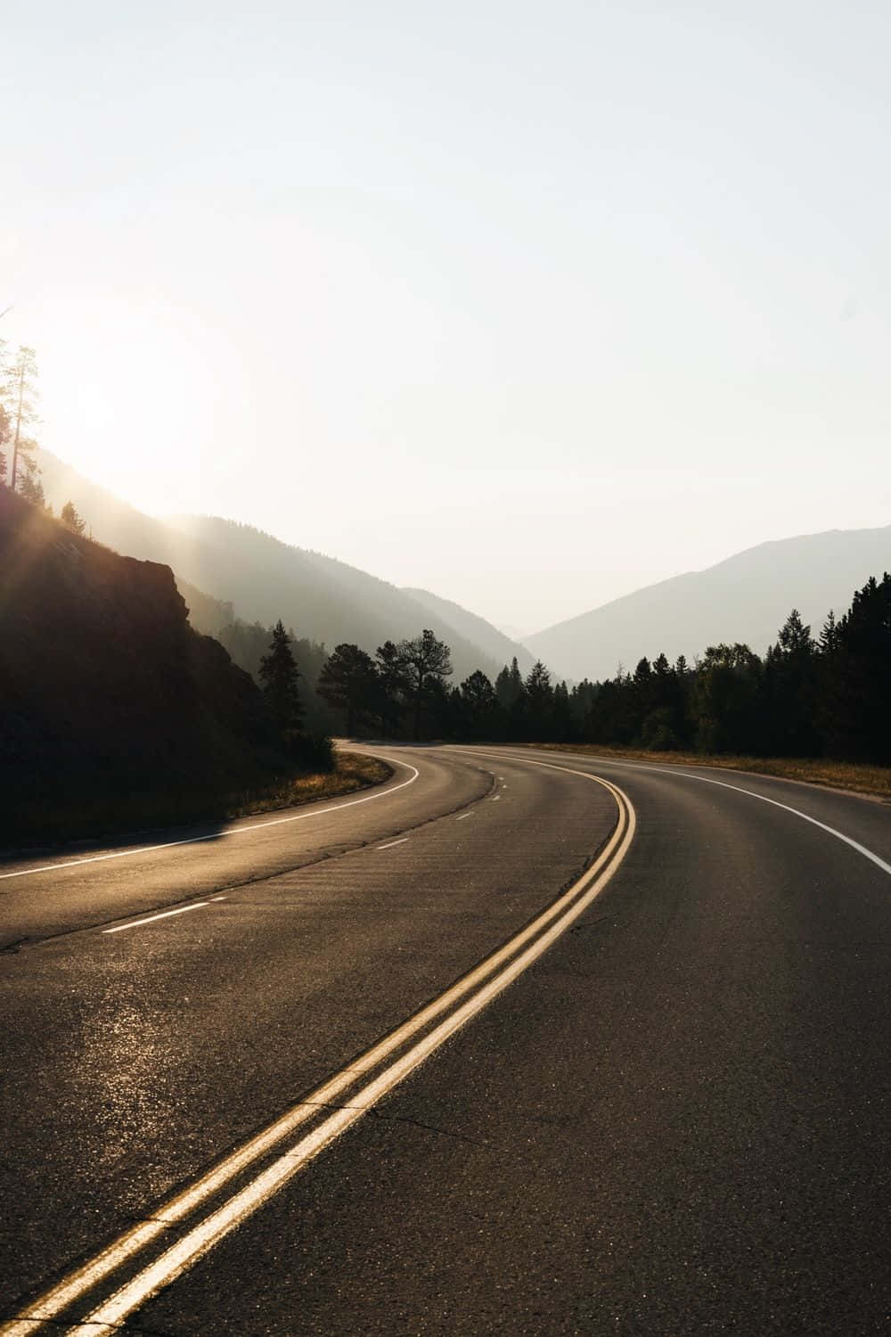 highway 桌布 壁紙 — 一條蜿蜒的瀝青公路在日出時分穿過霧氣繚繞的山巒，金色的光芒照亮了整個場景。