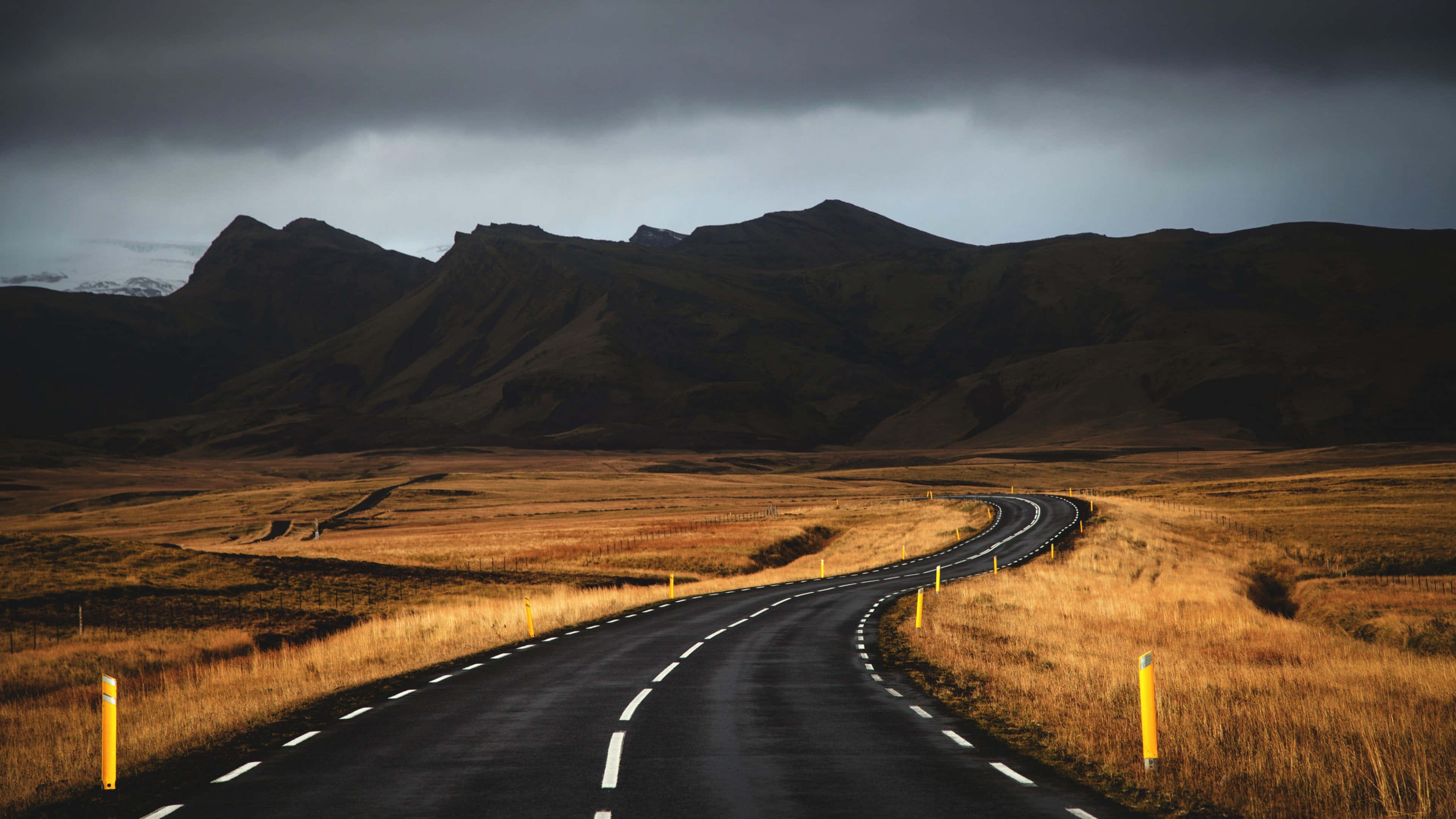 Iceland Desktop 桌布 壁紙 — 一條蜿蜒的瀝青路在戲劇性的多雲天空下，穿過金色的草原，遠處有黑暗的山脈。