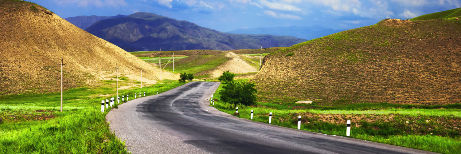 Country Road 桌布 壁紙 — 一條蜿蜒的瀝青路穿過充滿活力的綠色草原，兩旁是起伏的丘陵和遠處的山脈，在明亮的藍天下有散落的雲朵。