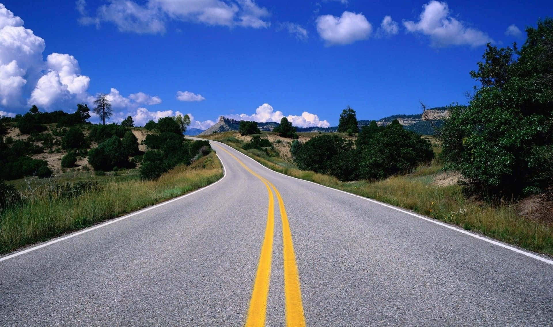 highway 桌布 壁紙 — 一條蜿蜒的瀝青道路，帶有黃色中心線，在陽光明媚的風景中蜿蜒而行，天空湛藍，點綴著蓬鬆的白雲。