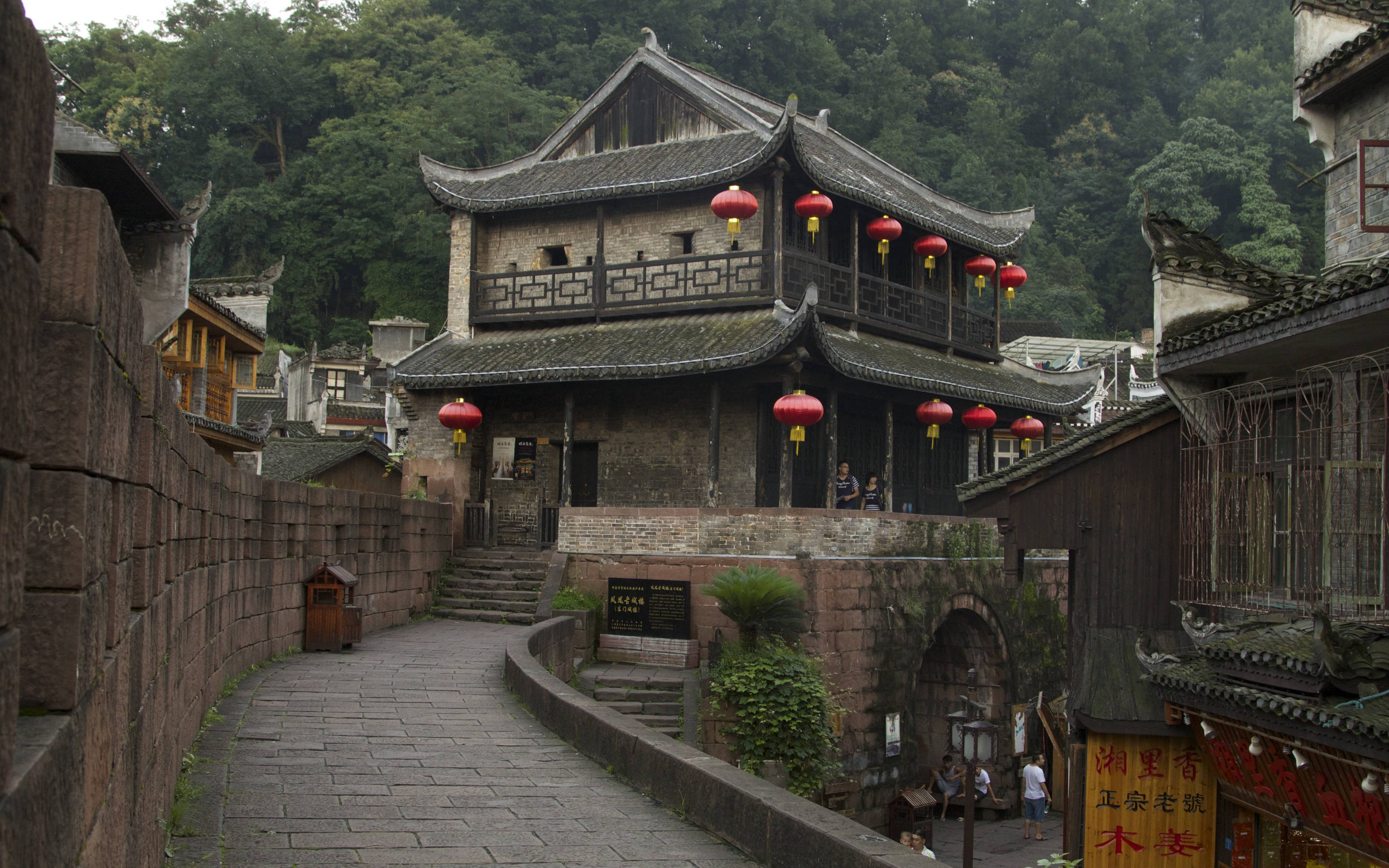 中式房屋壁紙 桌布 壁紙 — 一條蜿蜒的石徑穿過傳統中式建築，這些建築擁有瓦片屋頂和紅色燈籠，坐落在鬱鬱蔥蔥的綠色山丘之間。