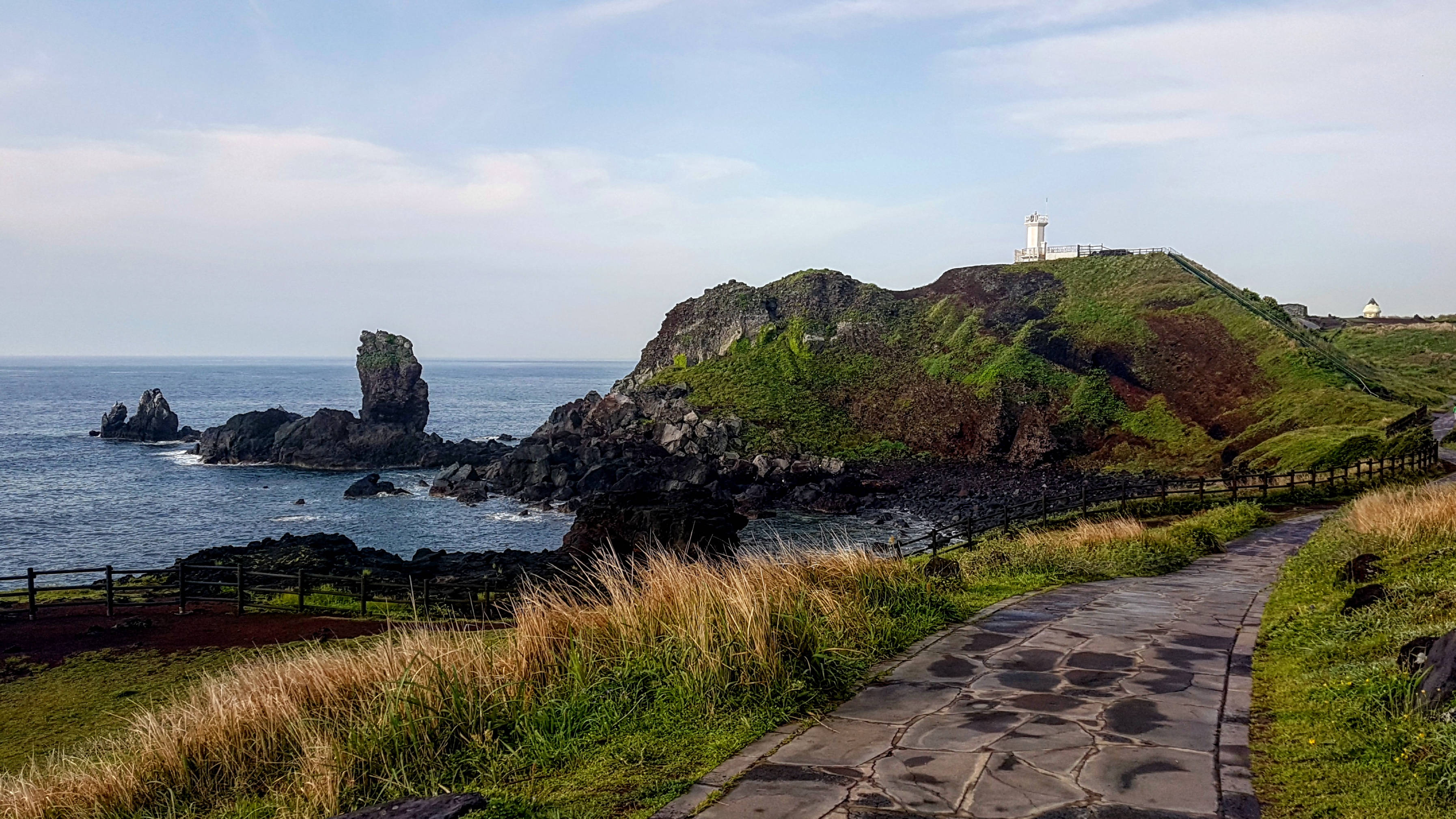 Jeju 桌布 壁紙 — 一條蜿蜒的石徑通往一座坐落在草叢懸崖上的燈塔，俯瞰著海洋和下方的岩石露頭。