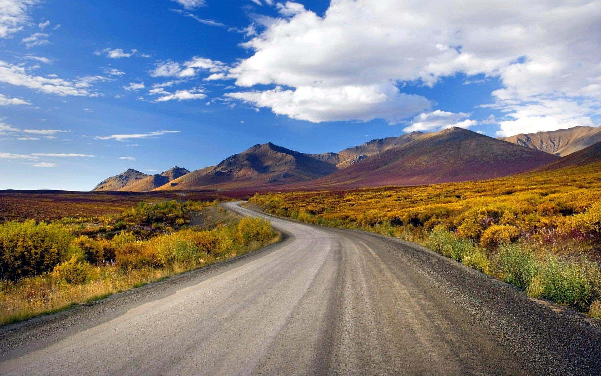 highway 桌布 壁紙 — 一條蜿蜒的碎石路穿過充滿活力的秋日景觀，上方是明亮的藍天和蓬鬆的白雲。