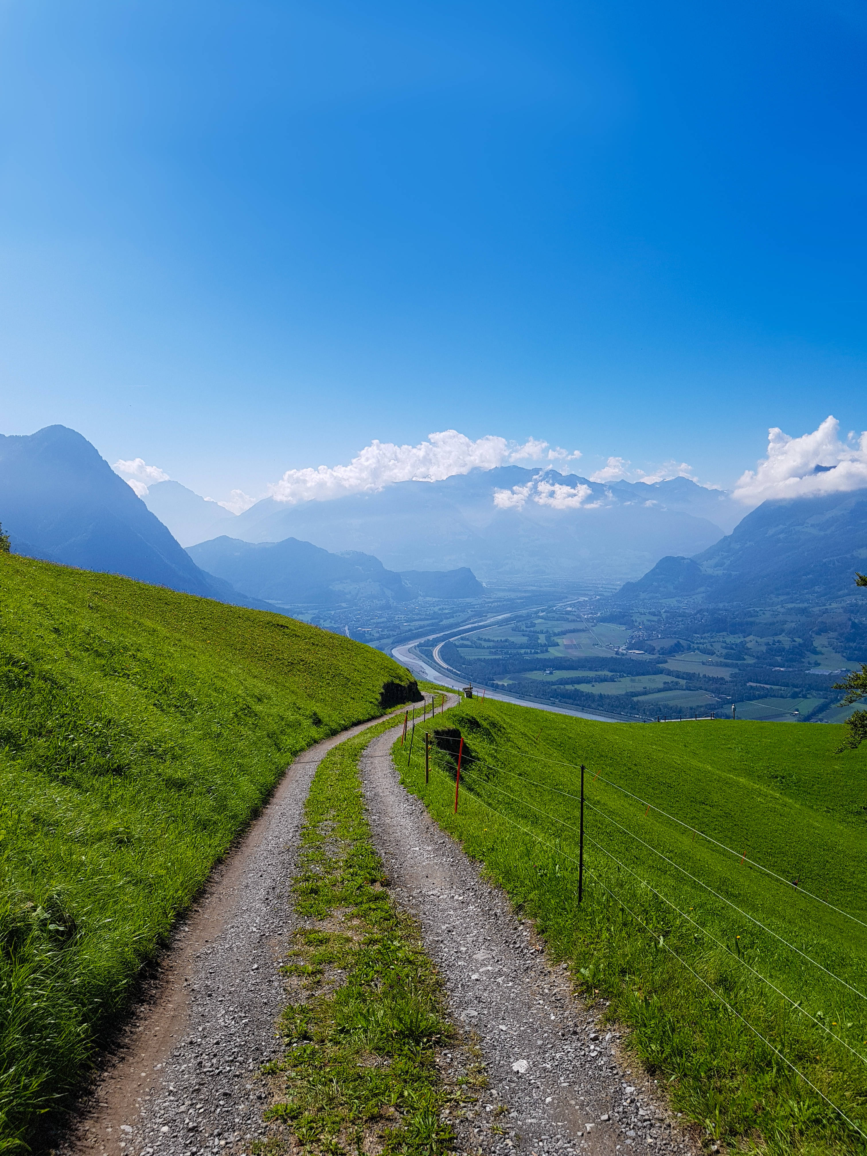 Liechtenstein 桌布 壁紙 — 一條蜿蜒的碎石路穿過充滿活力的綠色山丘，通向遠方的山脈和下方蜿蜒的河流，在晴朗的藍天下。