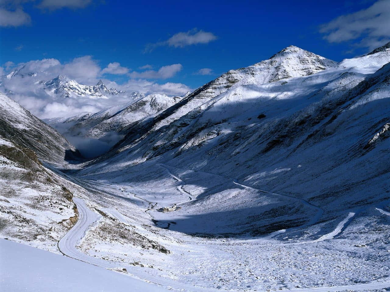 Beautiful Winter 桌布 壁紙 — 一條蜿蜒的積雪山路穿過戲劇性的阿爾卑斯山谷，上方是明亮的藍天和零星的白雲。