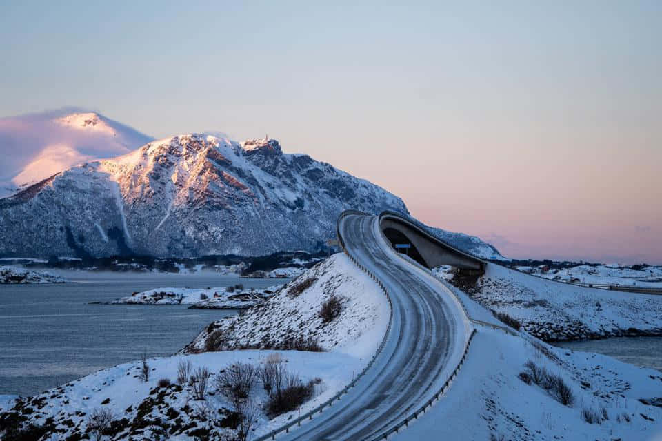 Road 桌布 壁紙 — 一條蜿蜒的積雪道路在暮色中戲劇性地彎曲越過峽灣，積雪覆蓋的山脈在柔和的粉紅色和藍色光芒中閃耀。
