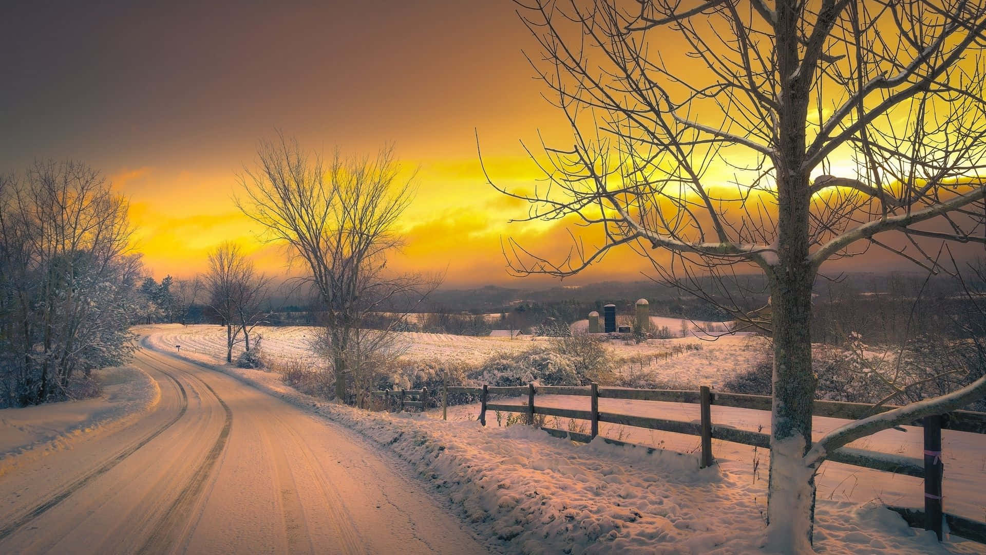 Winter Sunset 桌布 壁紙 — 一條蜿蜒的積雪道路穿過霜凍的冬季景觀，在充滿活力的橙色日落天空下。