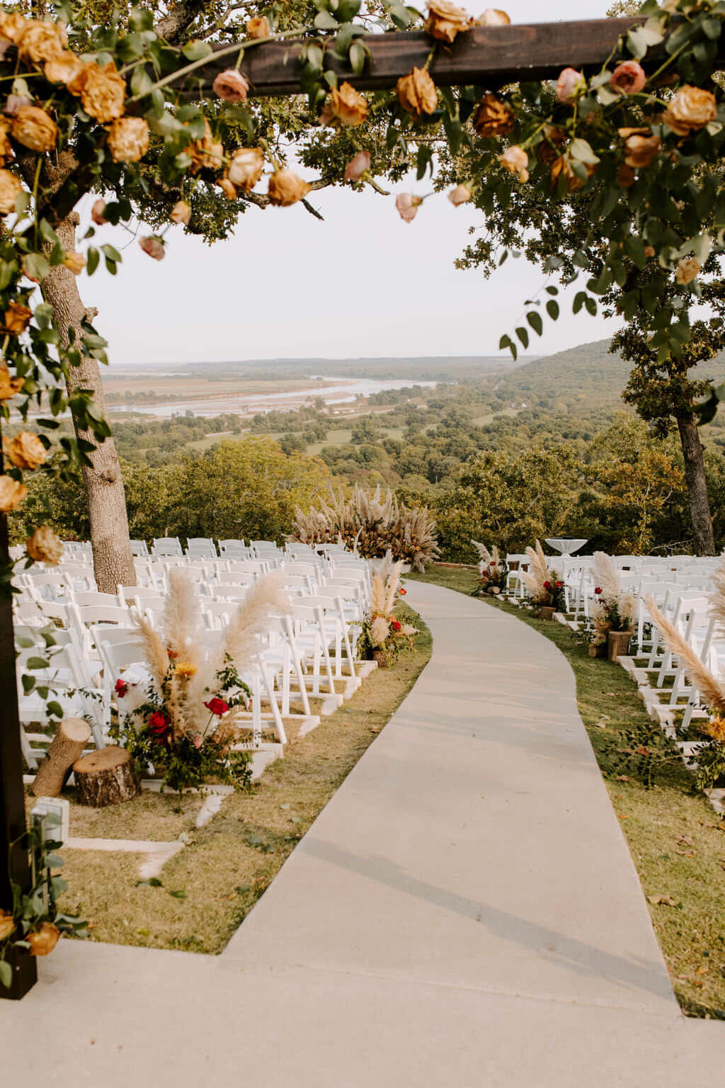 Wedding Aesthetic 桌布 壁紙 — 一條蜿蜒的通道，兩旁擺放著白色椅子和花卉佈置，通向一個裝飾著玫瑰的木製拱門，俯瞰著風景如畫的山谷。