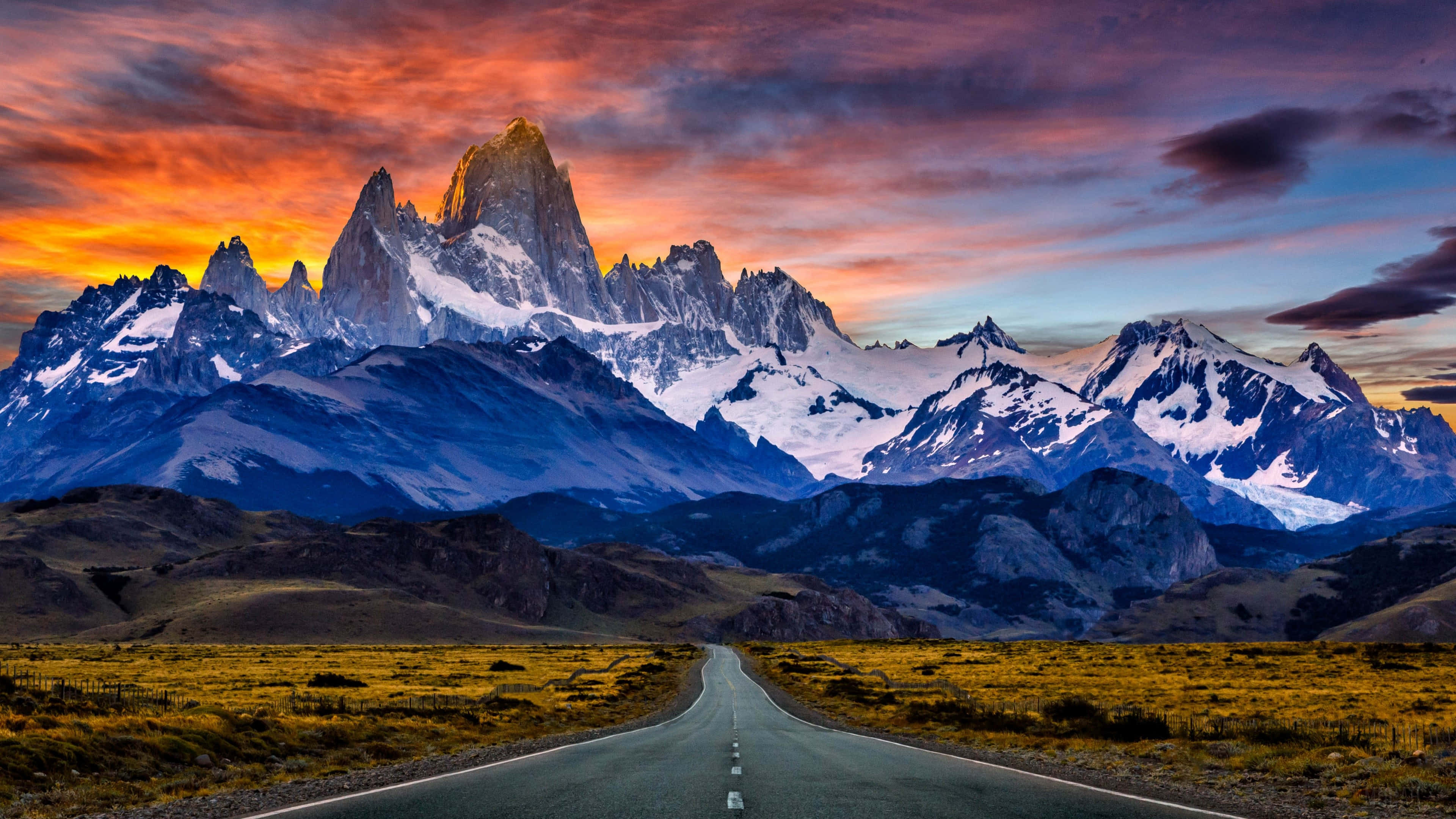 Patagonia 桌布 壁紙 — 一條蜿蜒的道路伸向白雪覆蓋的山峰，天空是火紅的夕陽，有鮮豔的橙色和紫色雲彩。
