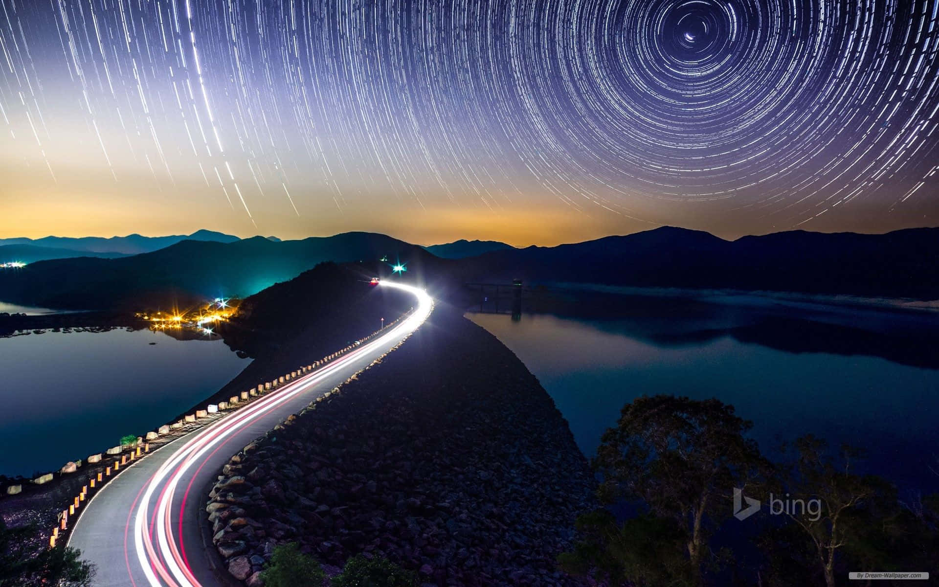 星軌在蜿蜒的道路上空盤旋，黃昏時水壩附近有汽車的光跡。 — 必應 壁紙