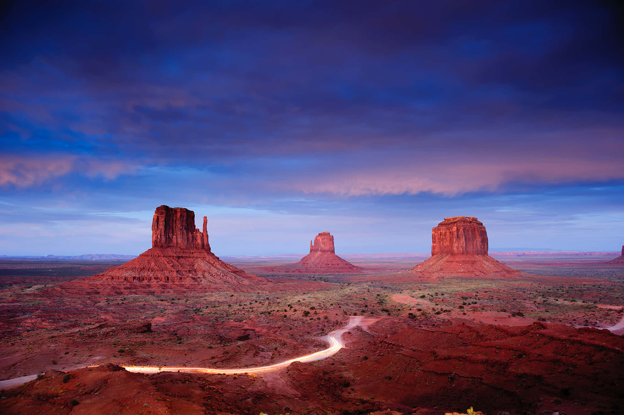 Arizona 桌布 壁紙 — 一條蜿蜒的道路在暮色中穿過沙漠景觀，三座巨大的紅色岩石在戲劇性的紫色和藍色天空下呈現剪影。