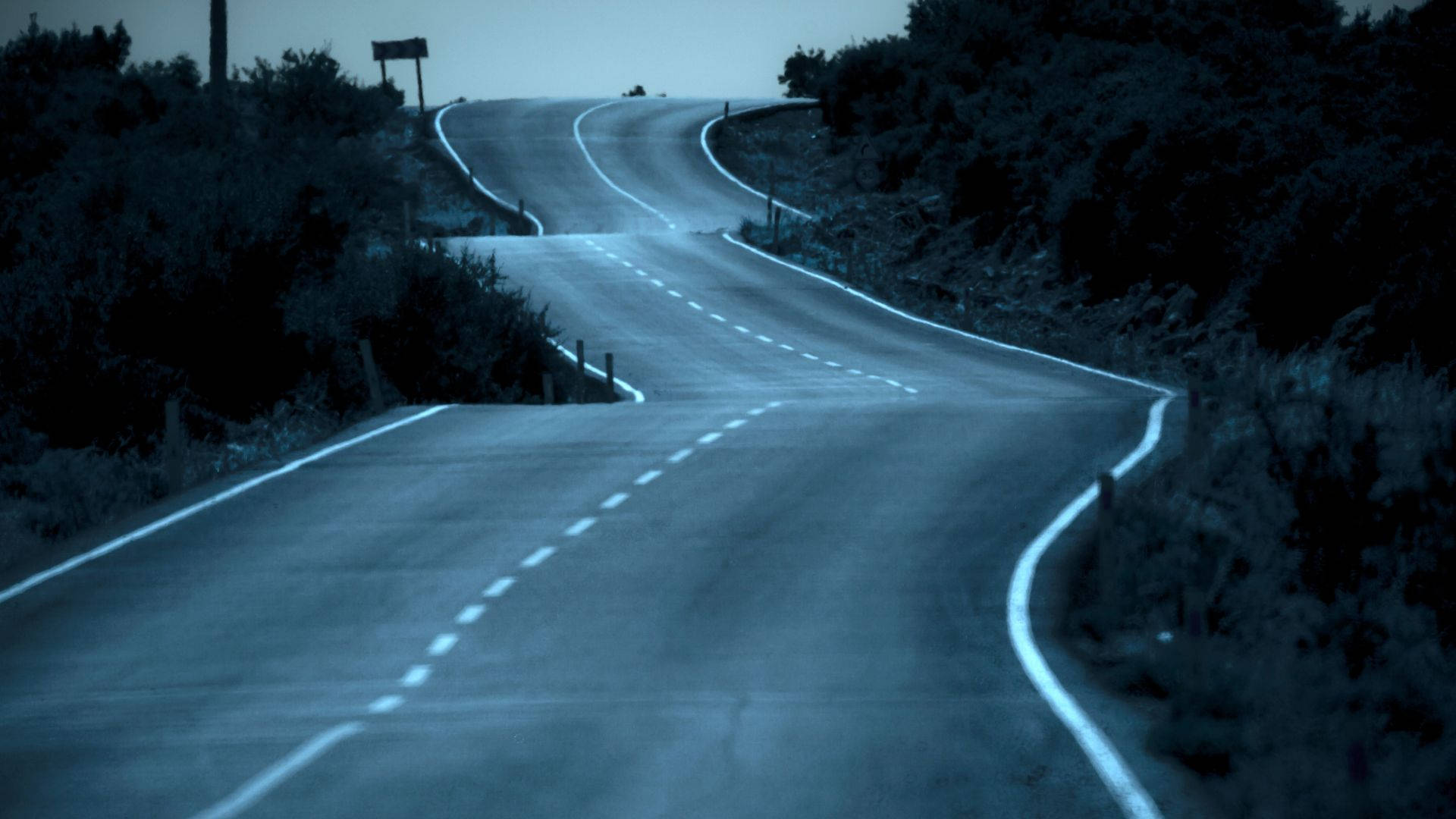 Road 桌布 壁紙 — 一條蜿蜒的道路在暮色天空下彎曲進入迷霧遠方，兩旁是黑暗的灌木叢和發光的白色線條。
