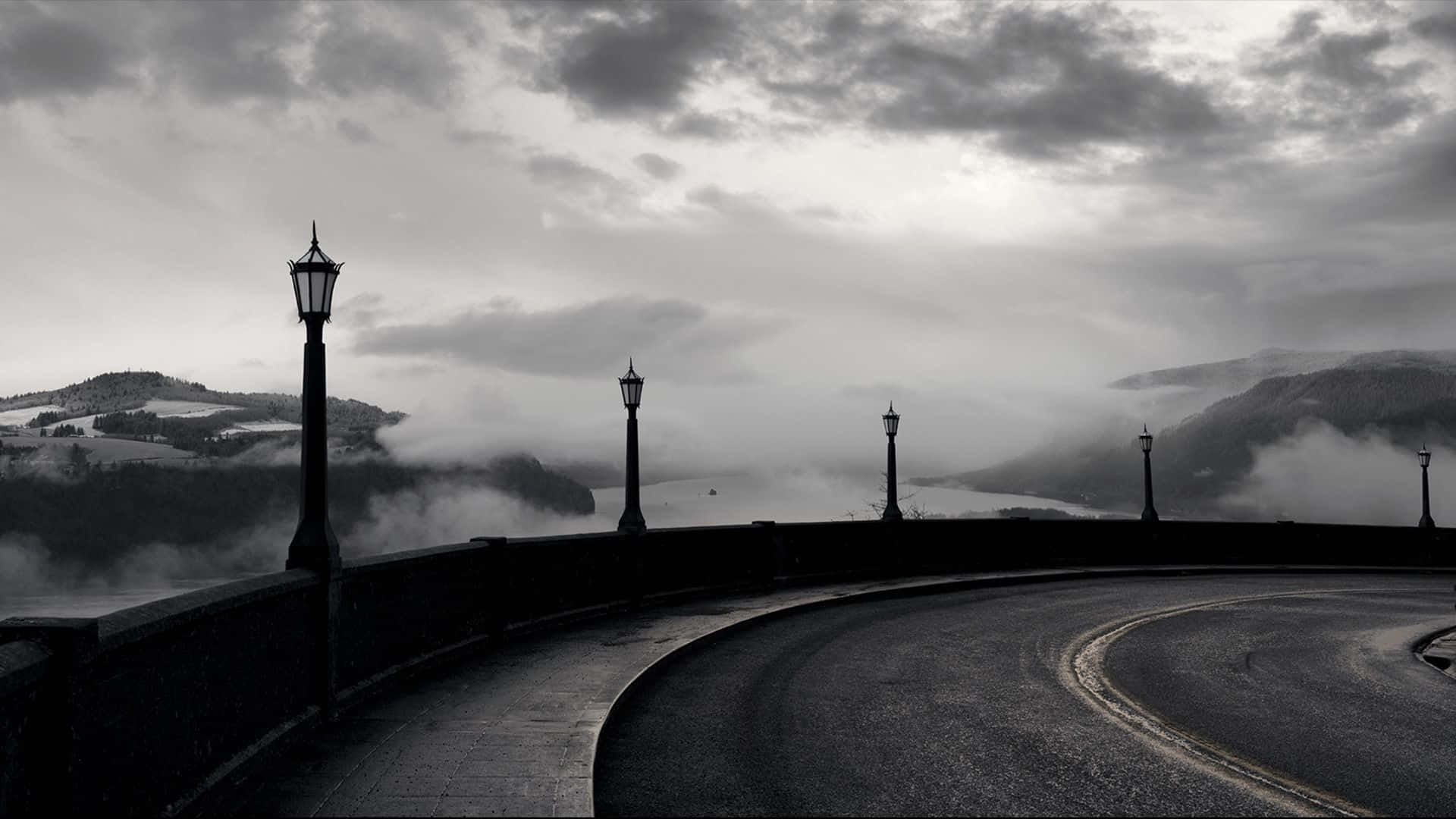 黑白 桌布 壁紙 — 一條蜿蜒的道路在濃雲密佈的天空下，穿過霧氣瀰漫的山谷，兩旁有復古路燈。