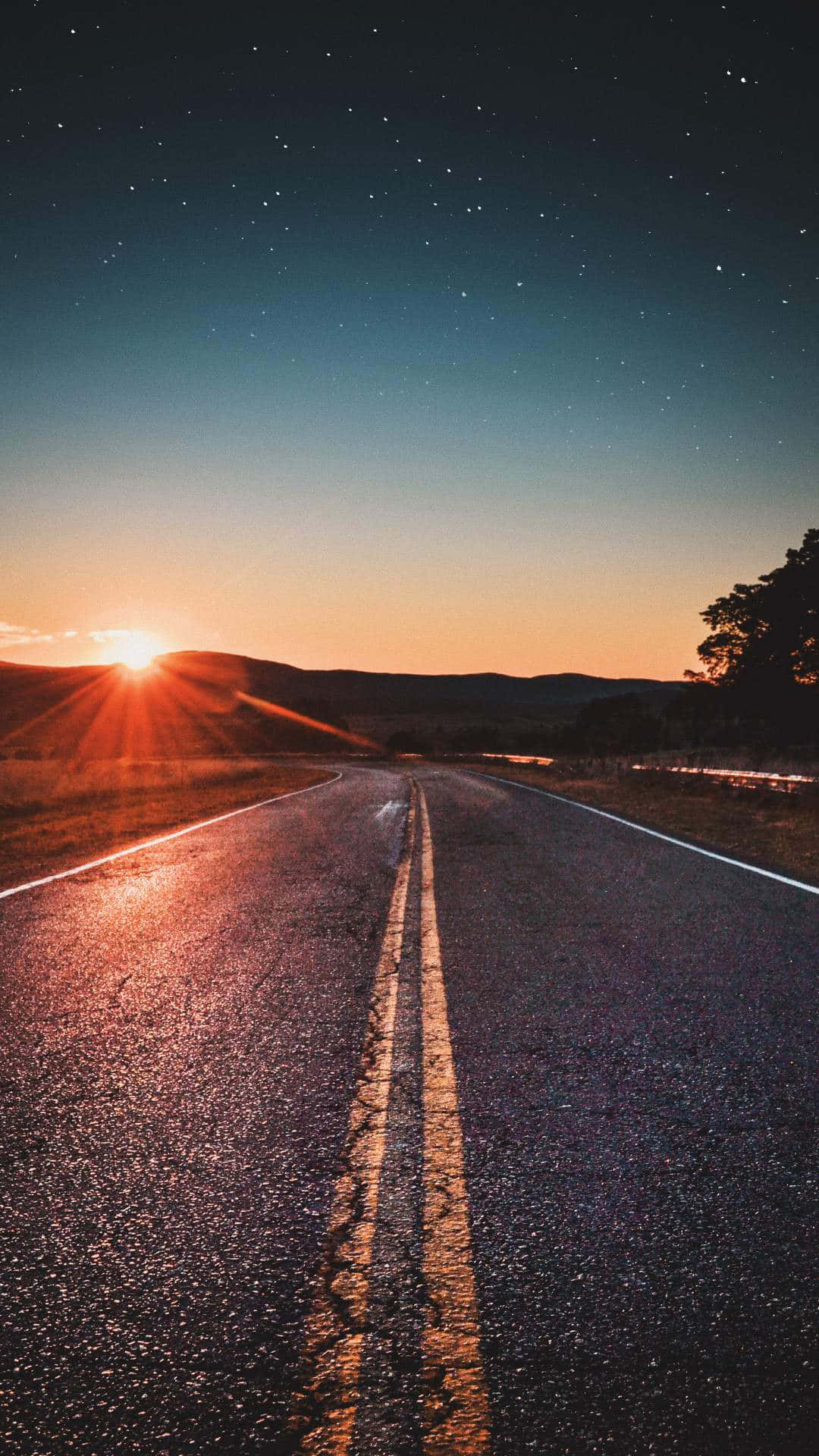 Sunrise iPhone 桌布 壁紙 — 一條蜿蜒的道路在繁星點點的暮色天空下延伸至地平線，地平線上有發光的太陽。
