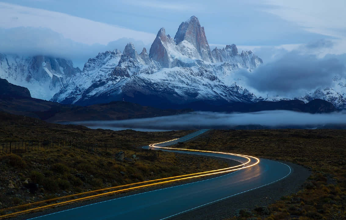 Patagonia 桌布 壁紙 — 一條蜿蜒的道路在黃昏時分彎向被雲霧部分遮蓋的雪山，帶有發光的汽車軌跡。