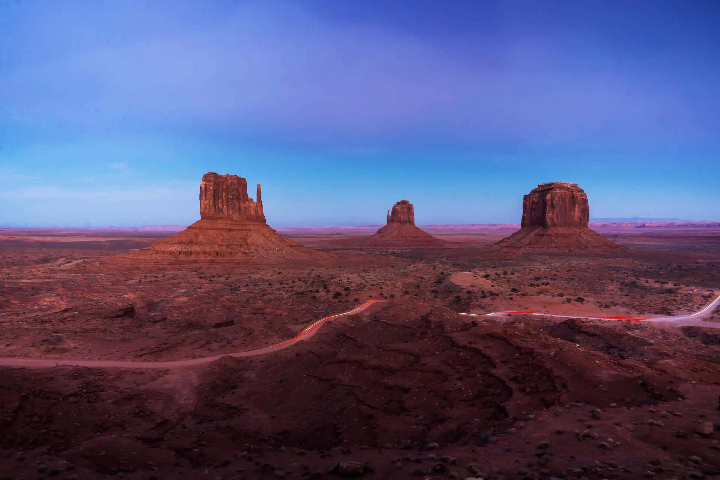 Arizona 桌布 壁紙 — 一條蜿蜒的道路在黃昏時分穿過廣闊的沙漠景觀，三座標誌性的孤峰在漸變的藍色天空下呈現剪影。