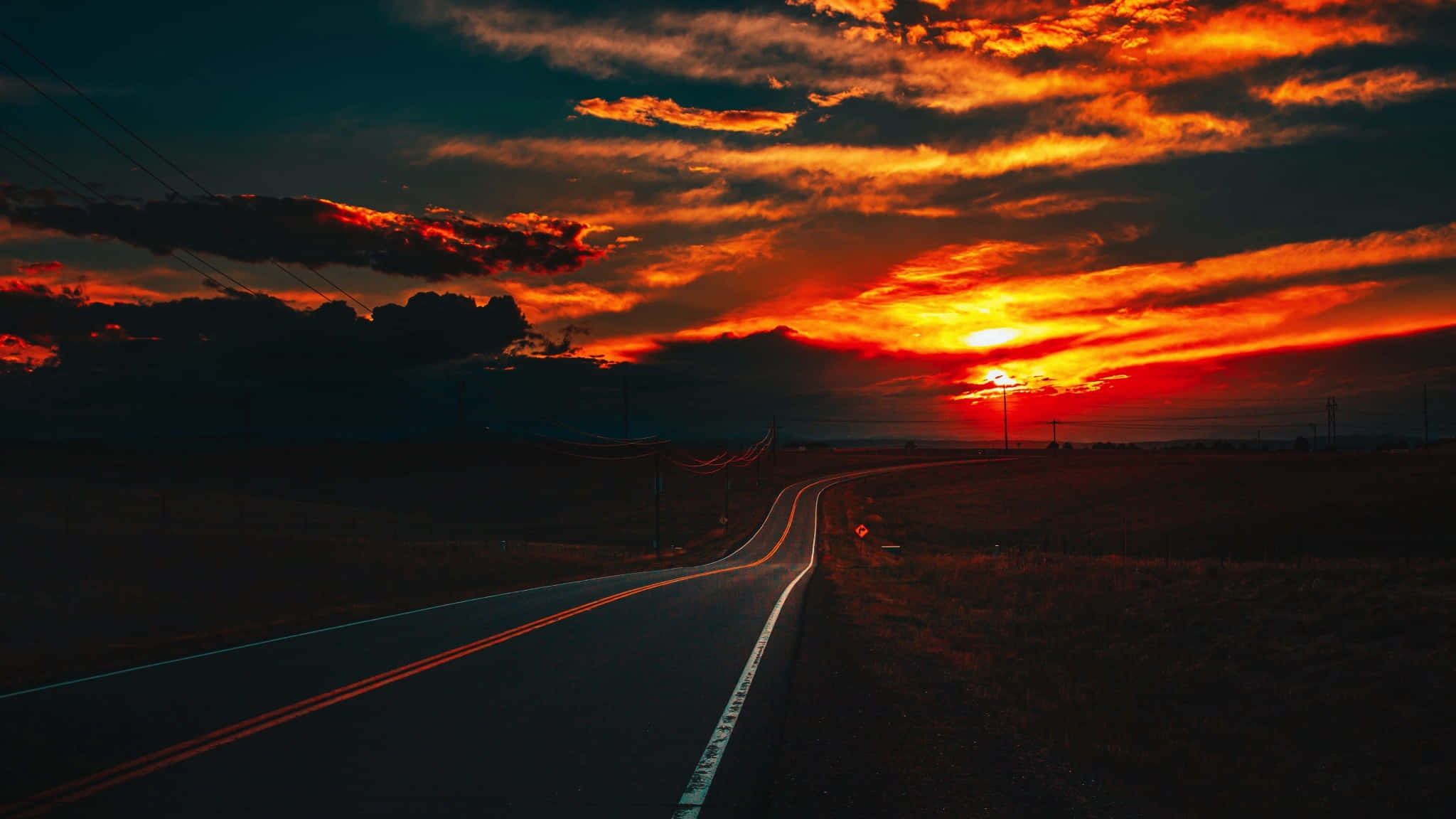 2048x1152 桌布 壁紙 — 一條蜿蜒的道路延伸至火紅的日落，伴隨著橙色的雲朵和黑暗的輪廓山丘。