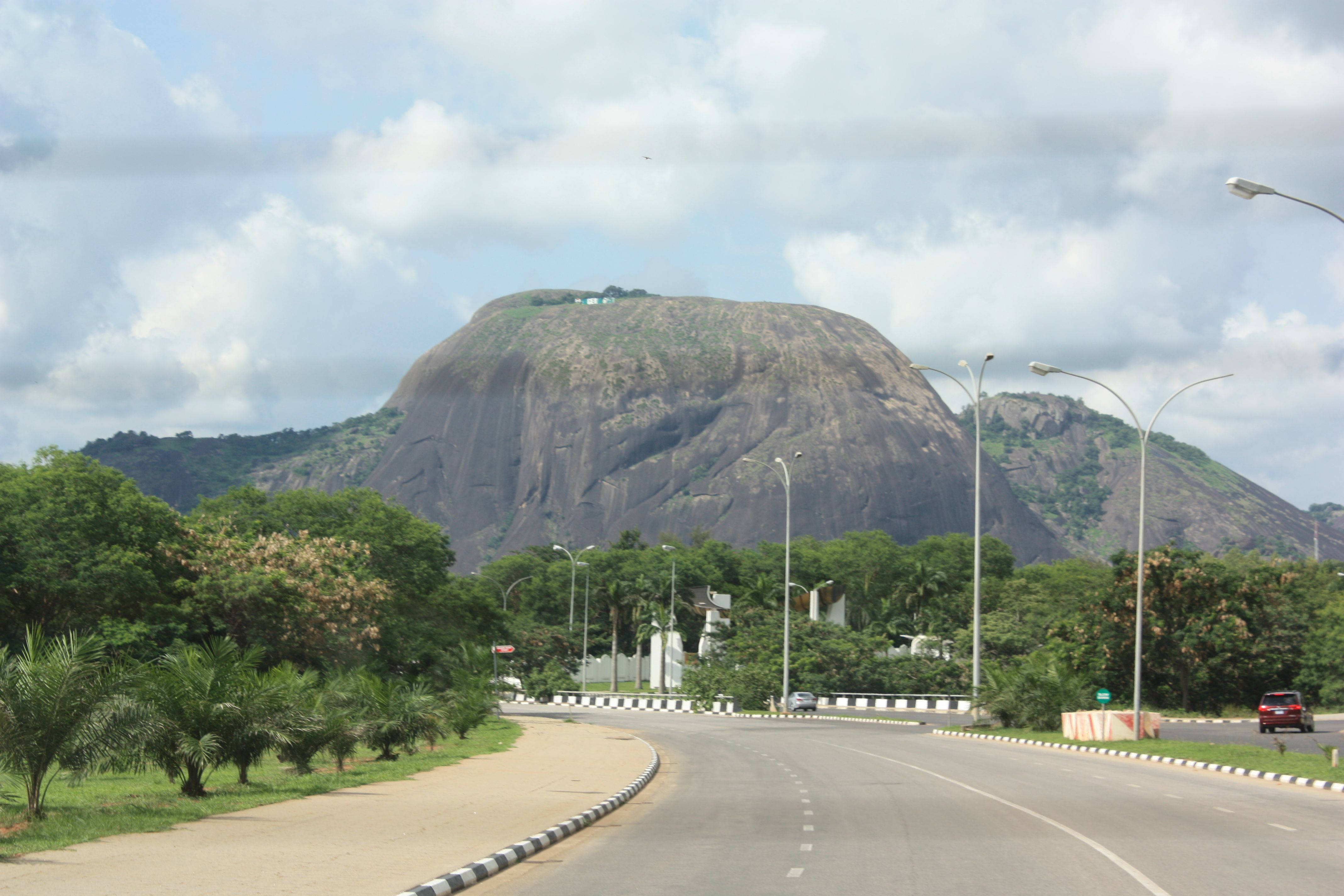 Nigeria 桌布 壁紙 — 一條蜿蜒的道路彎向一座巨大的岩石山丘，山頂覆蓋著綠植，天空多雲。