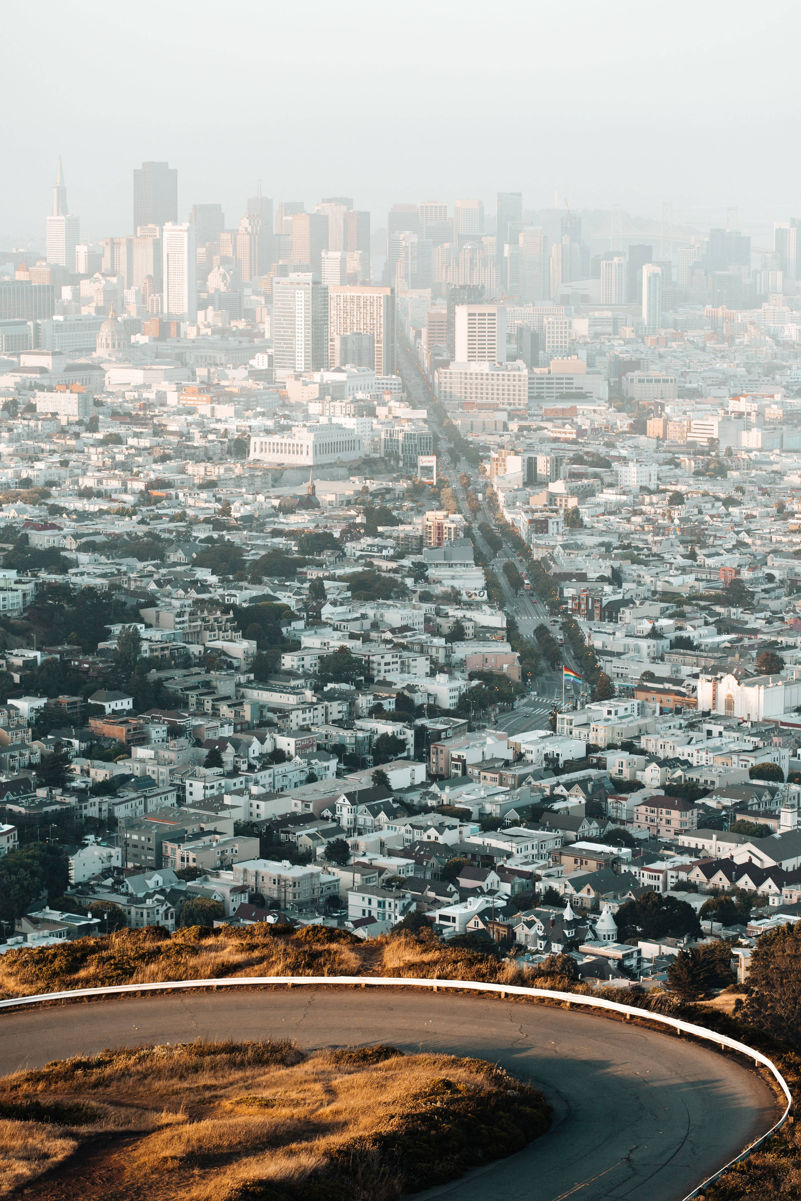 San Francisco Phone 桌布 壁紙 — 一條蜿蜒的道路沿著山坡向下延伸，通往煙霧瀰漫的舊金山天際線，城市密集擴展，遠處高樓林立。