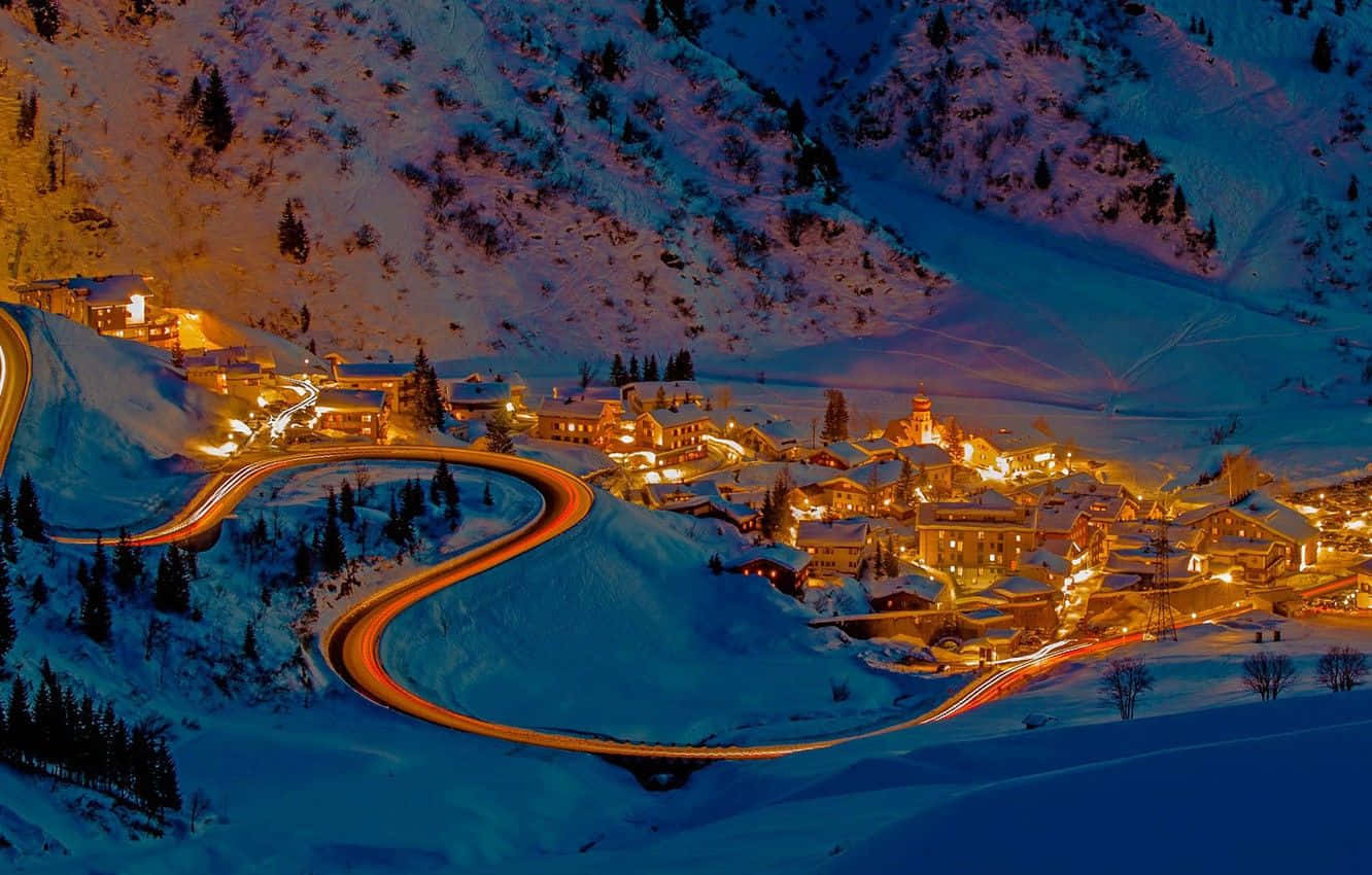 Ski Resort 桌布 壁紙 — 一條蜿蜒的道路穿過一個被雪覆蓋的山村，夜晚閃爍著溫暖的燈光。