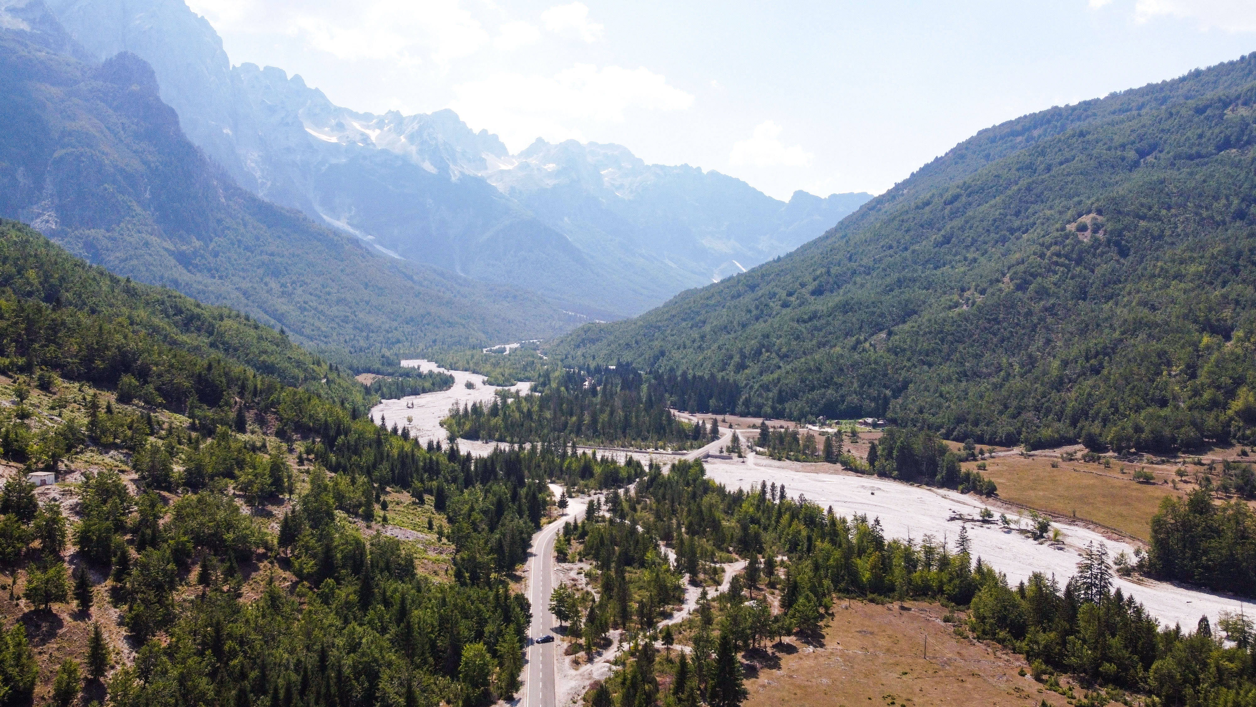 Albania 桌布 壁紙 — 一條蜿蜒的道路穿過一個鬱鬱蔥蔥的綠色山谷，兩旁是高聳的森林覆蓋的山脈，山頂積雪，天空明亮。
