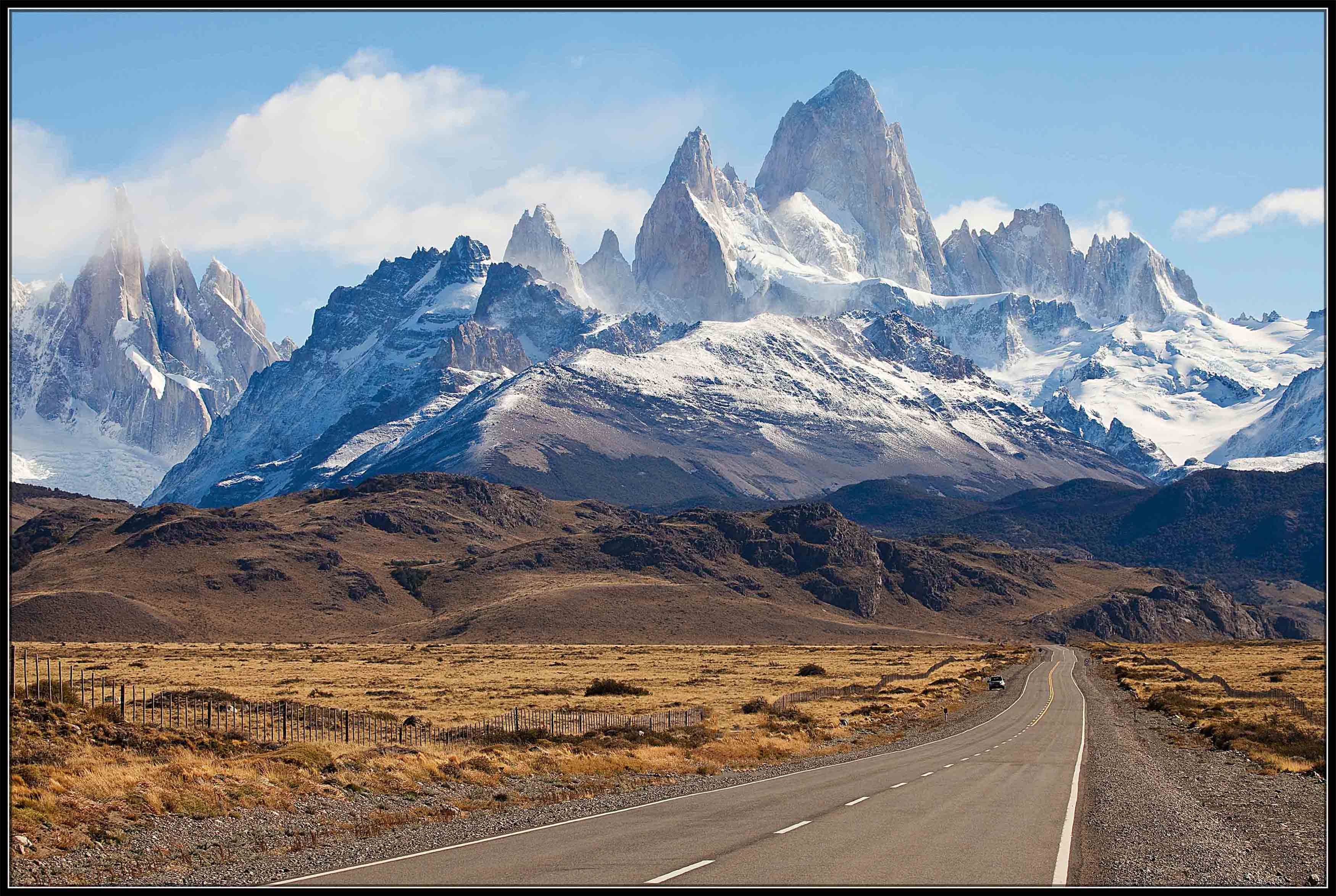 Patagonia 桌布 壁紙 — 一條蜿蜒的道路穿過乾旱的平原，通往高聳的雪山頂峰，天空湛藍，雲朵稀疏。
