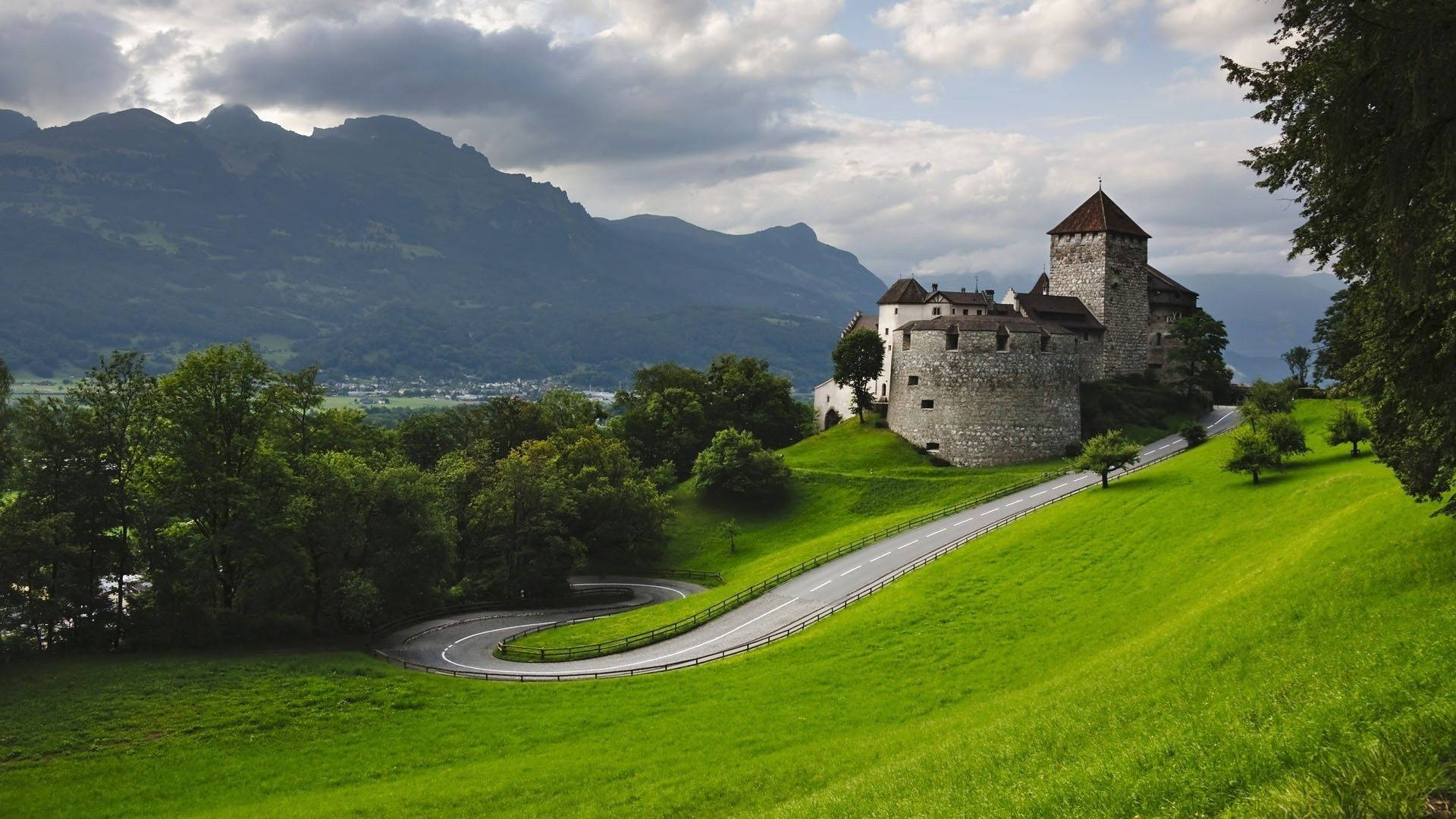 Liechtenstein 桌布 壁紙 — 一條蜿蜒的道路穿過充滿活力的綠色山丘，通往一座歷史悠久的石頭城堡，城堡坐落在山頂上，背景是山脈，天空多雲。