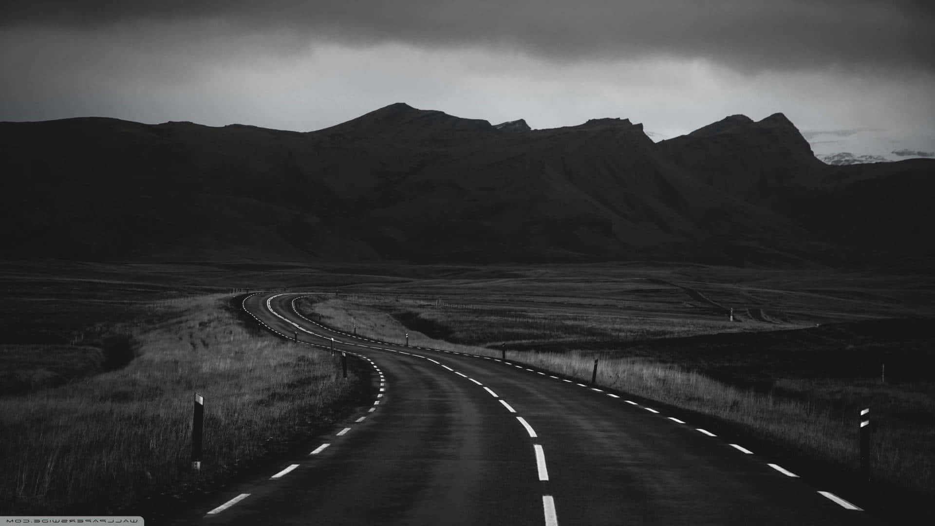 Black And White Aesthetic Pc 桌布 壁紙 — 一條蜿蜒的道路穿過單色調的荒涼景觀，天空陰沉，遠山可見。