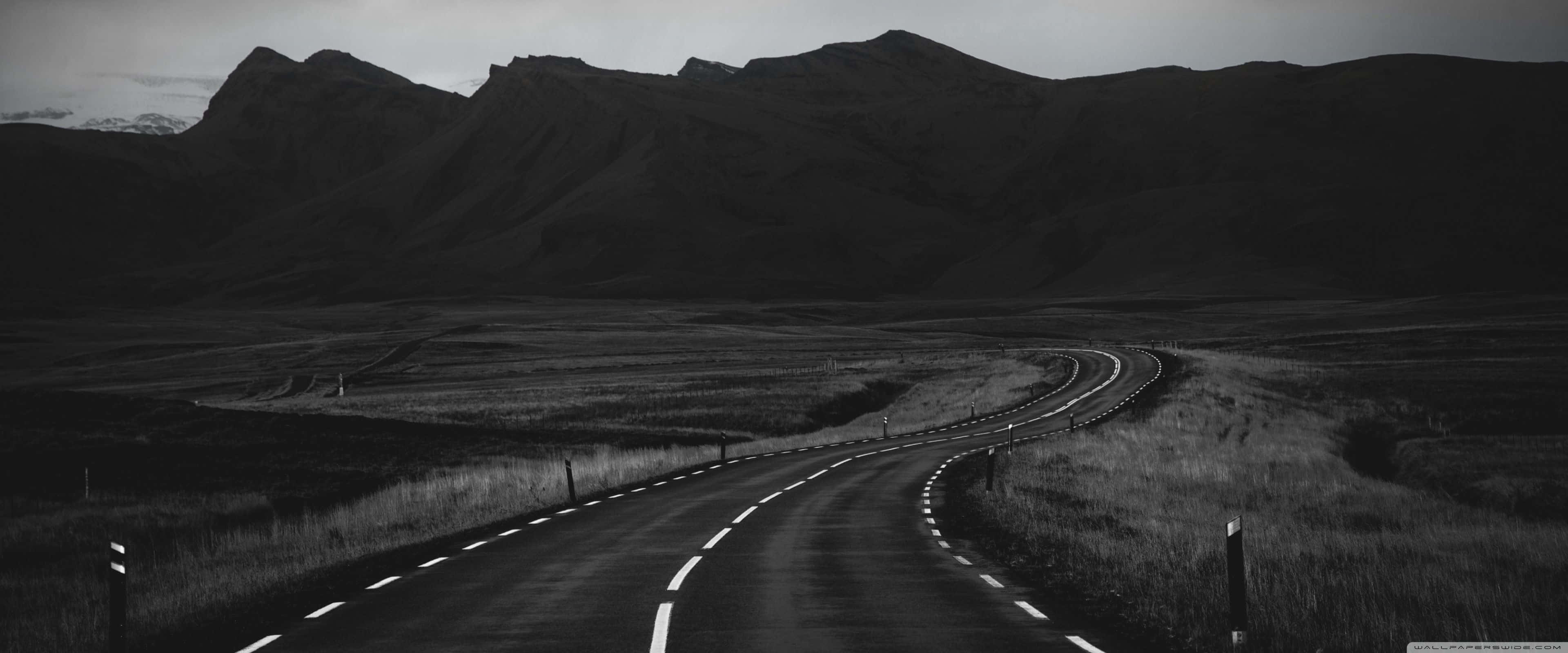 Apple 3840x1600 桌布 壁紙 — 一條蜿蜒的道路穿過單調的黑白景觀，遠處有鋸齒狀的山脈聳立。