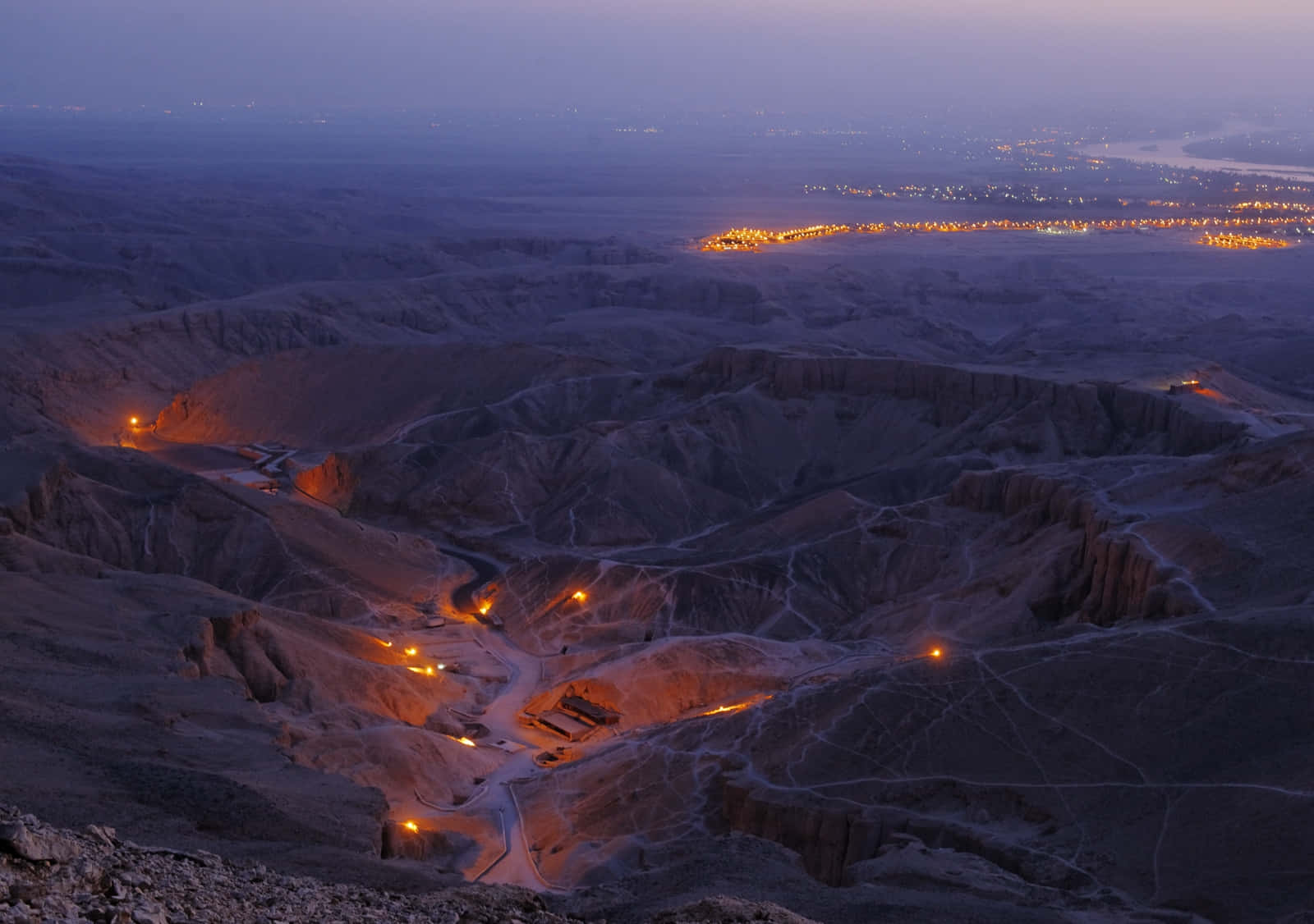 Valley Of The Kings 桌布 壁紙 — 一條蜿蜒的道路穿過崎嶇的沙漠峽谷，在黃昏時分閃爍著溫暖的橙色燈光，通往遠處的城市景觀。