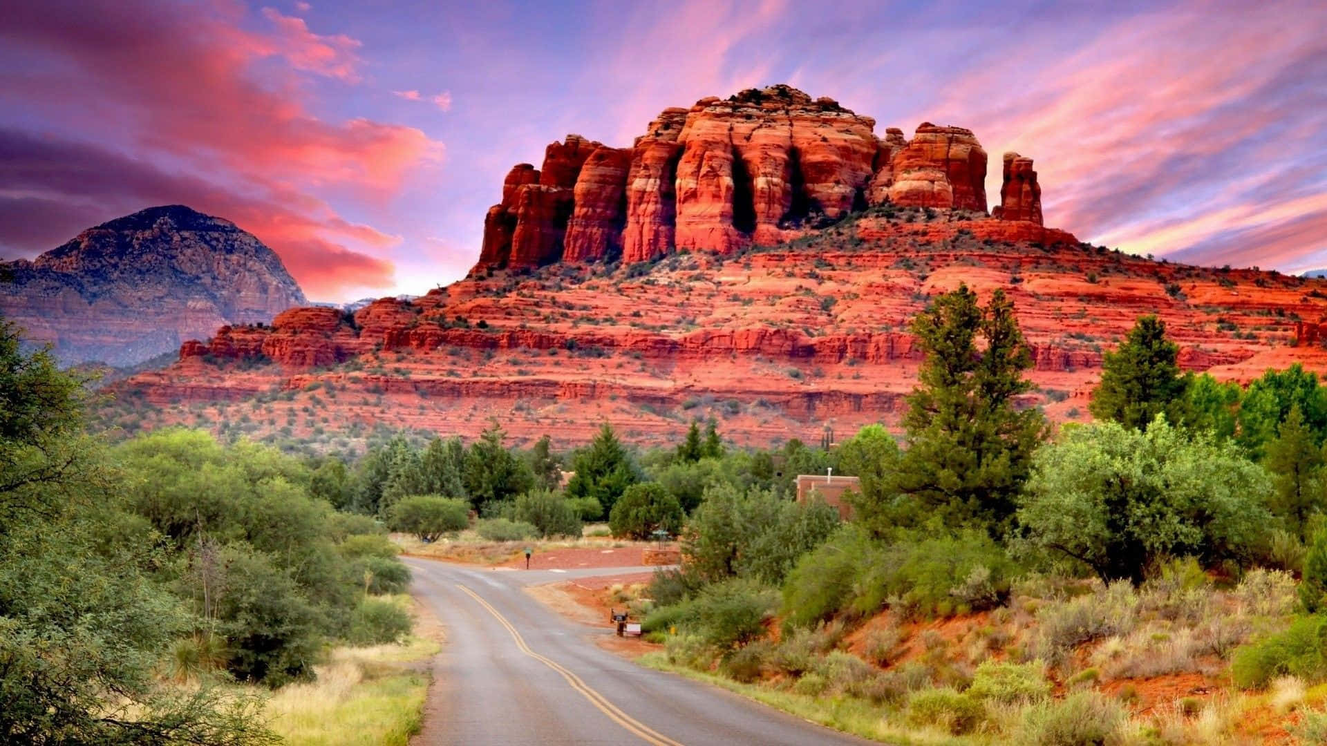 Arizona 桌布 壁紙 — 一條蜿蜒的道路穿過沙漠景觀，通向高聳的紅色岩層，天空呈現出充滿活力的粉紅色和紫色日落。