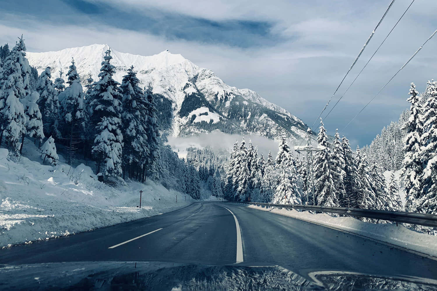 雪山 桌布 壁紙 — 一條蜿蜒的道路穿過白雪覆蓋的森林，雪覆蓋的山脈在陰雲密布的天空下聳立。