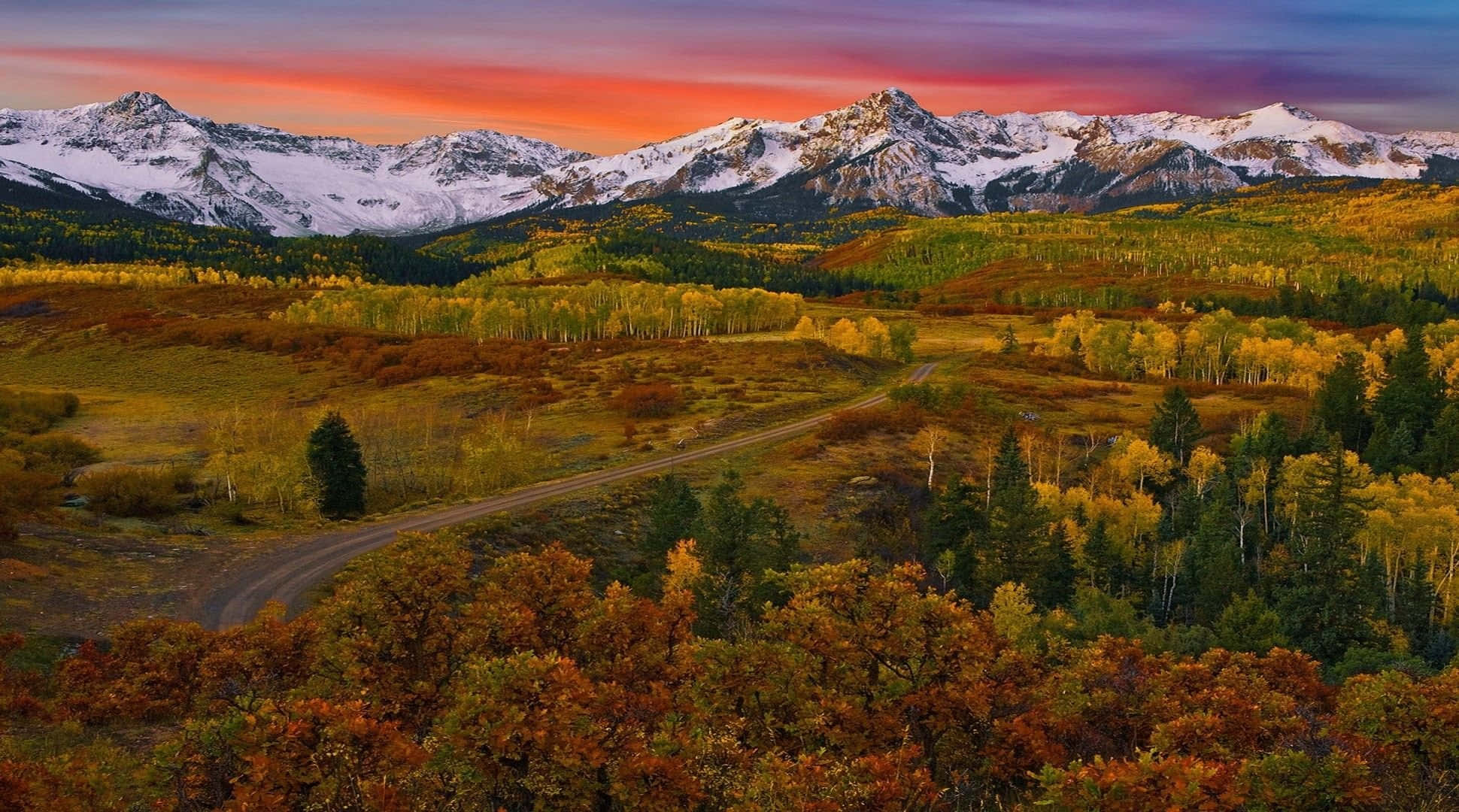 Colorado Mountains 桌布 壁紙 — 一條蜿蜒的道路穿過秋天的森林，通往在熾熱日落天空下的雪山。