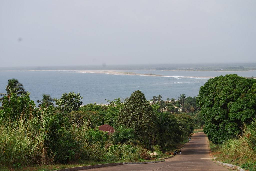 Liberia 桌布 壁紙 — 一條蜿蜒的道路穿過茂密的綠色樹葉，通向遠處的海灘和陰天下的平靜藍色大海。