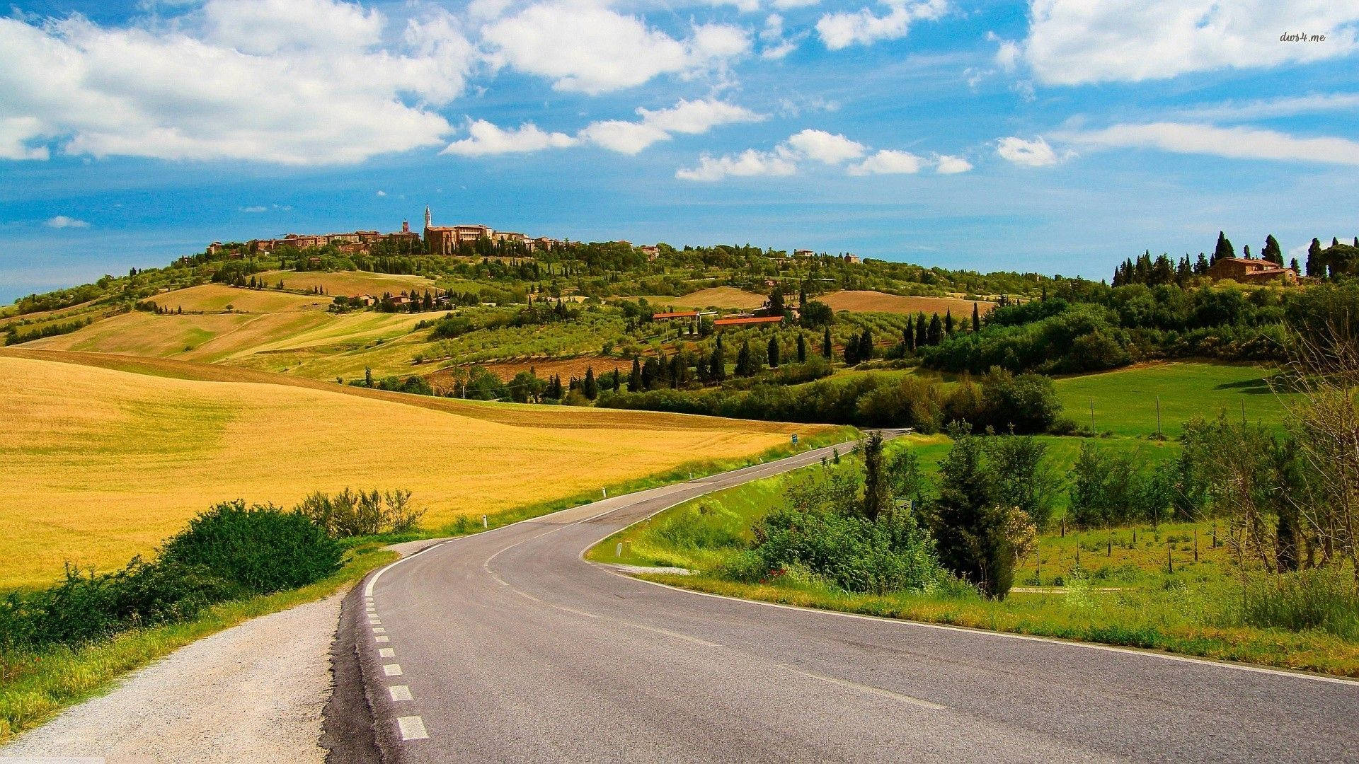 Country Road 桌布 壁紙 — 一條蜿蜒的道路穿過金色和綠色的托斯卡納山丘，通往一個山頂村莊，上方是明亮的藍天和蓬鬆的白雲。