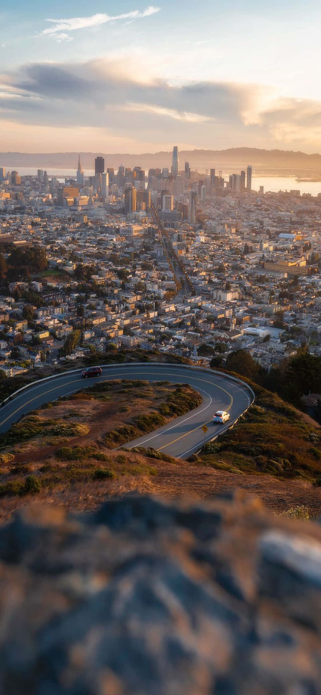 iPhone X California 桌布 壁紙 — 一條蜿蜒的道路穿過金色的山丘，俯瞰沐浴在溫暖夕陽下的舊金山天際線。