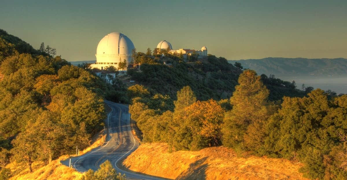 Observatory 桌布 壁紙 — 一條蜿蜒的道路穿過金黃色的秋樹，向上延伸至山頂上白色圓頂的天文台，正值日出時分。
