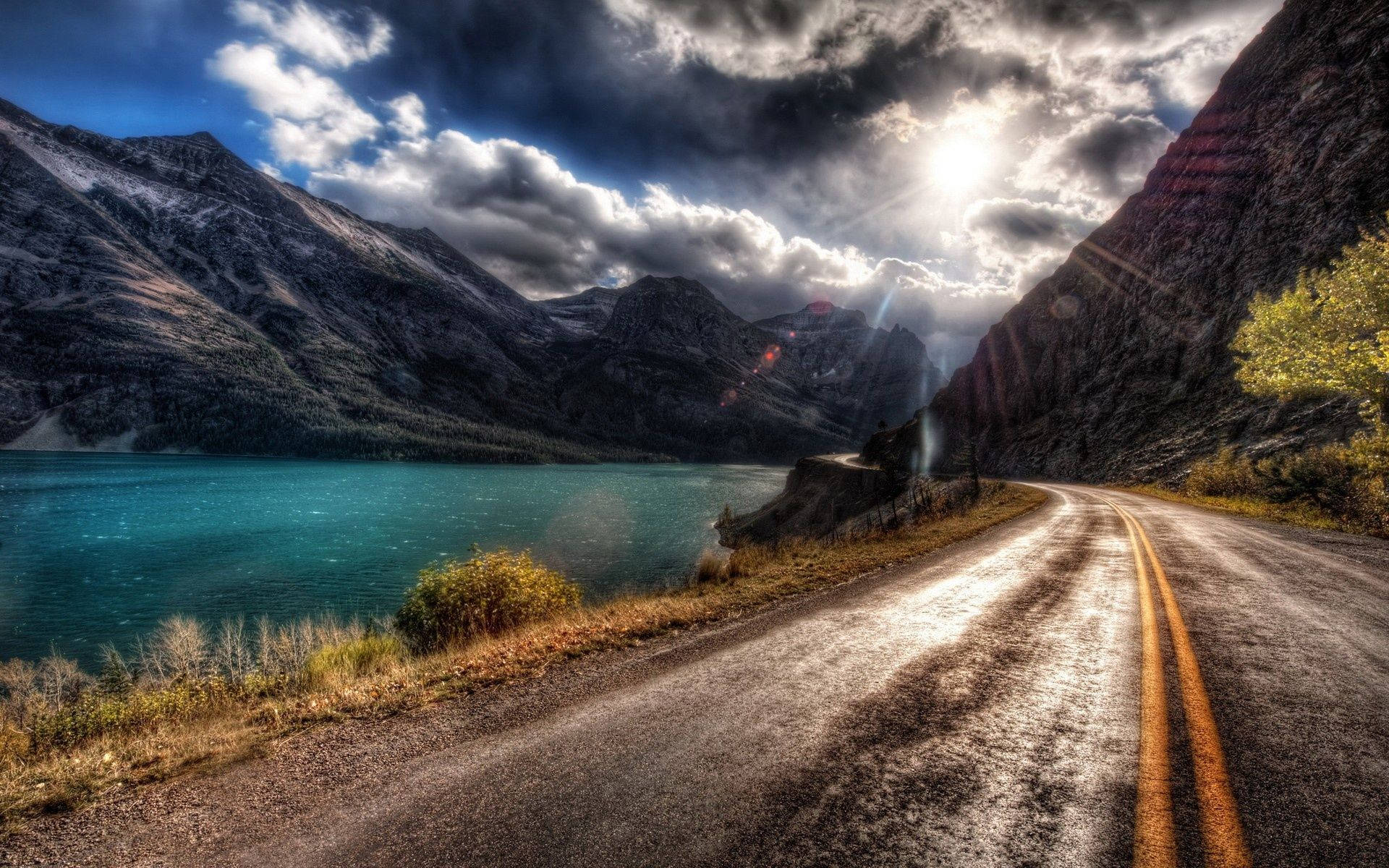 山景 桌布 壁紙 — 一條蜿蜒的道路緊貼著一個碧綠色的湖泊，旁邊是白雪覆蓋的山脈，天空戲劇性地多雲，陽光穿透雲層。